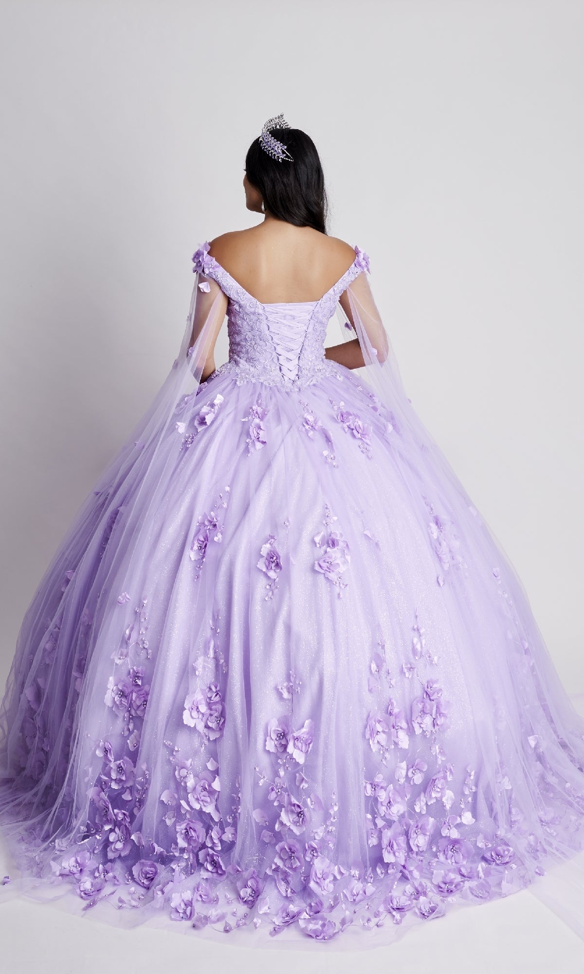 A woman in the Nina Canacci Q100 long quinceanera dress, featuring off-the-shoulder floral lace appliqués and a tiara, stands facing away against a plain white background.
