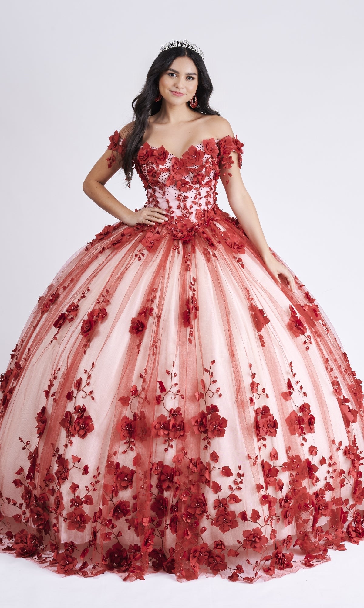A young woman smiles in the Nina Canacci Q102 long Quinceanera dress, a red and pink off-the-shoulder ball gown with 3D floral appliques, posing against a plain white background and wearing a tiara.