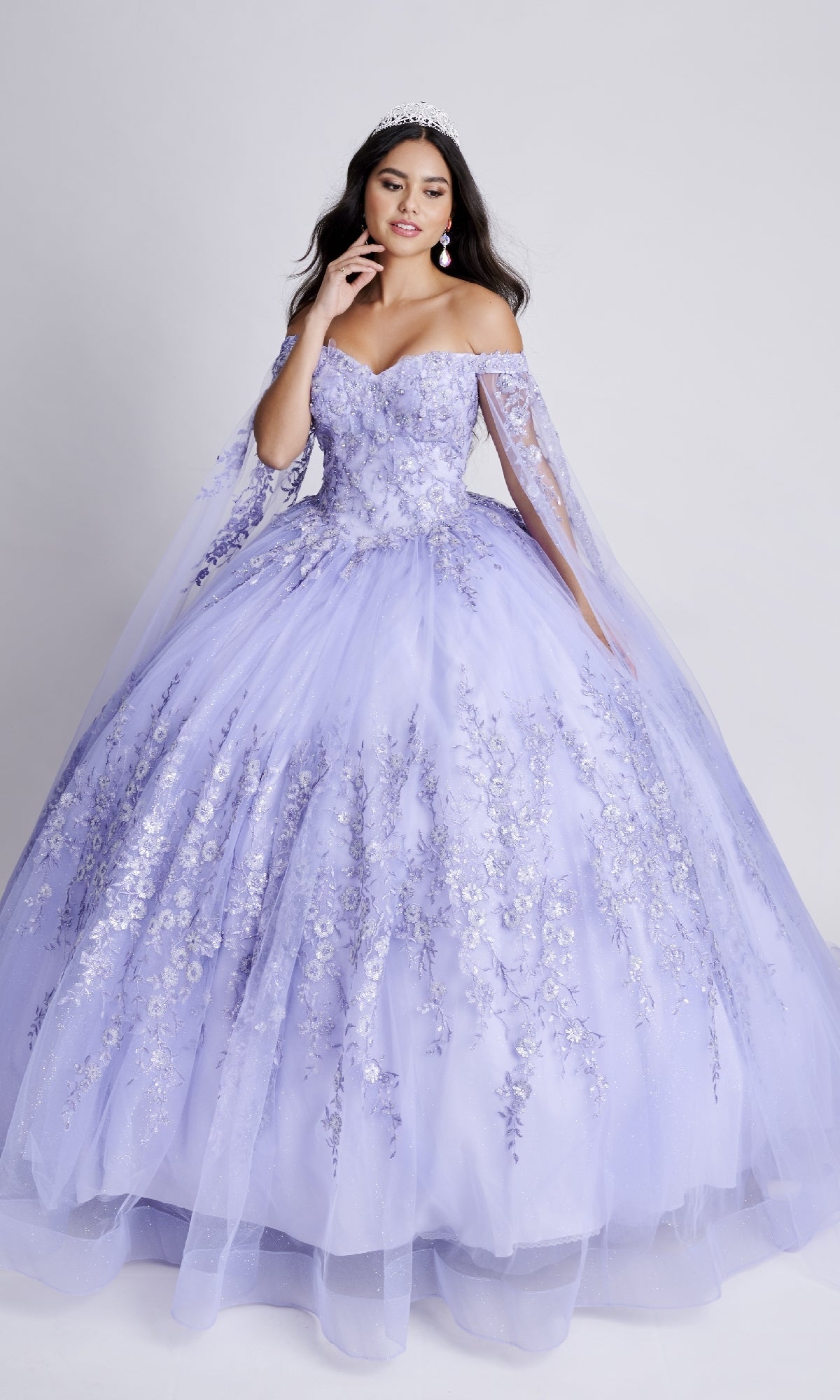 Model in Nina Canacci Q109 long quinceanera dress, a lavender ball gown with floral embroidery and off-the-shoulder sleeves, wearing a tiara, posed against a plain light background.