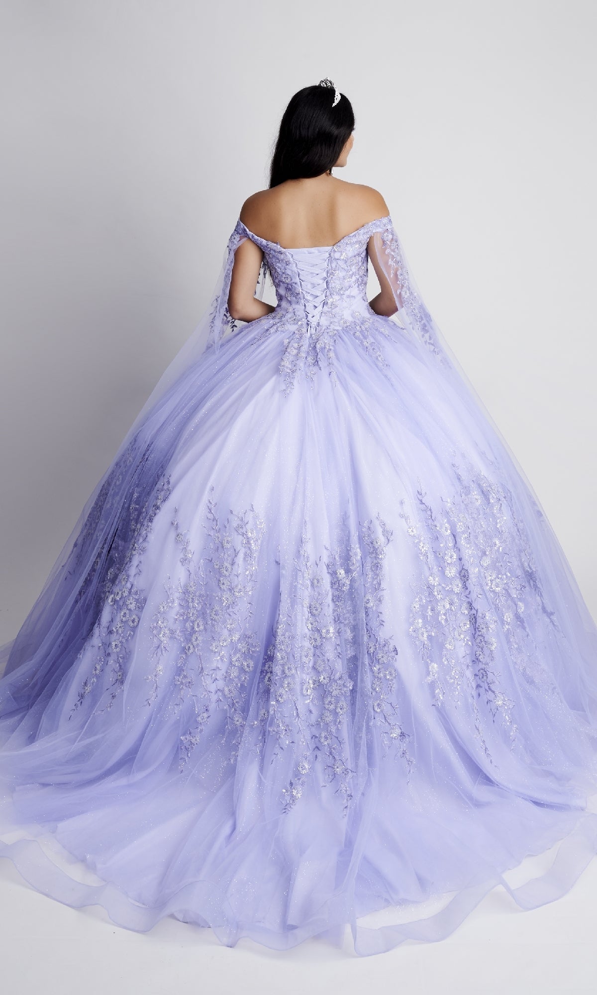 A woman with long dark hair models the Nina Canacci Q109 Long Quinceanera Dress, an off-the-shoulder gown with a floral bodice and full lavender tulle skirt, pictured from the back against a plain background.