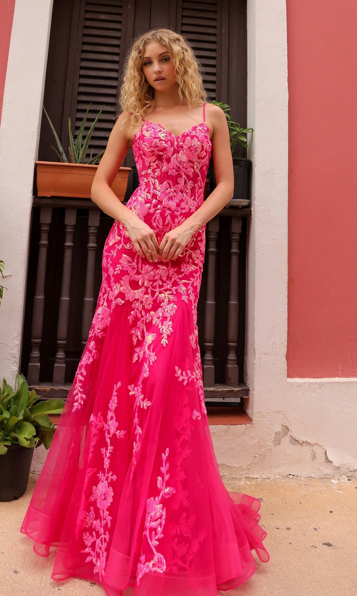 A woman stands by a shuttered window wearing the Sequin-Lace Long Neon Mermaid Prom Dress Q1390, featuring spaghetti straps, floral designs, and a fitted, flared silhouette.