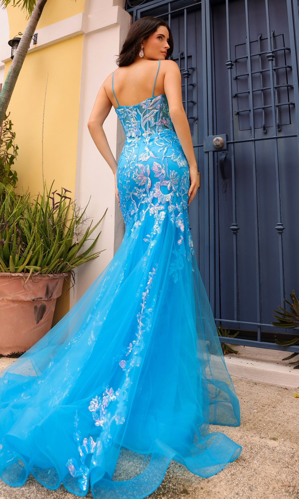 A woman stands outdoors by a blue door, wearing the Sequin-Lace Long Neon Mermaid Prom Dress Q1390, featuring floral embroidery and a tulle train.