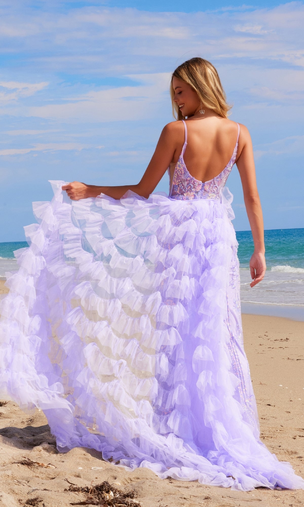 A woman stands on a sandy beach under a blue sky, facing the ocean with her back to the camera, wearing the Nox Anabel Q1542 Ruffled Long Beaded Prom Dress with a removable ruffled overskirt in lavender.