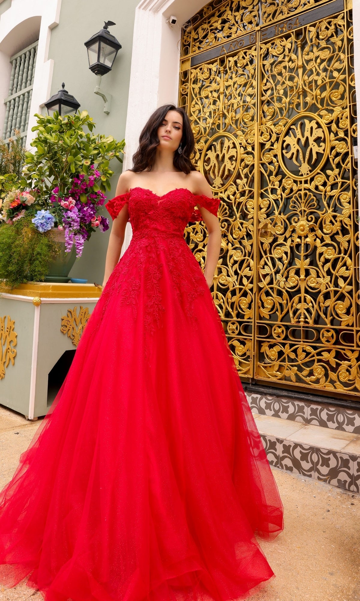 A woman in the Off-the-Shoulder Long Tulle Prom Ball Gown R1303 stands before ornate gold doors and a pale green building adorned with flower arrangements.