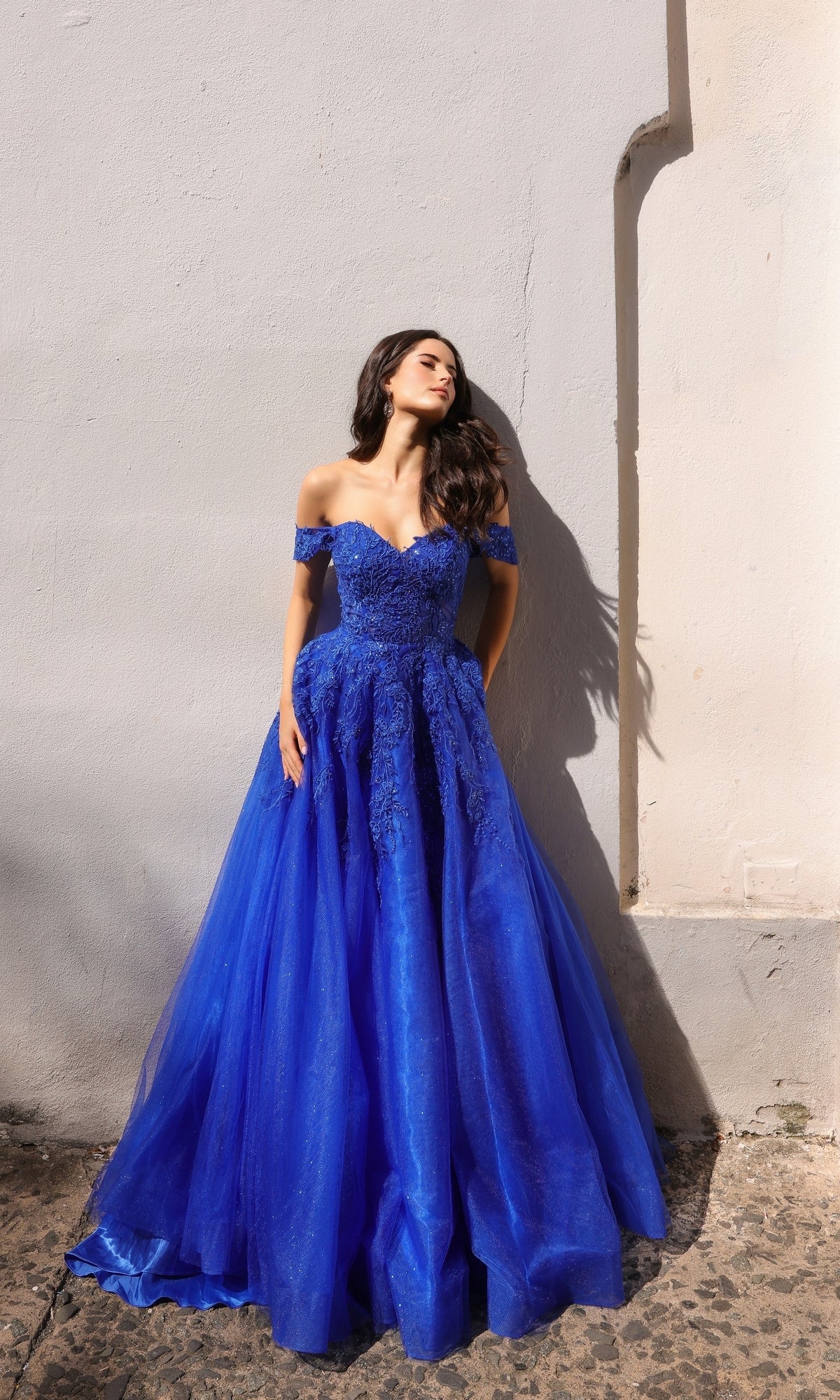 A woman wearing the Off-the-Shoulder Long Tulle Prom Ball Gown R1303 in bright blue stands against a light-colored wall, looking to the side with one hand resting on her dress.