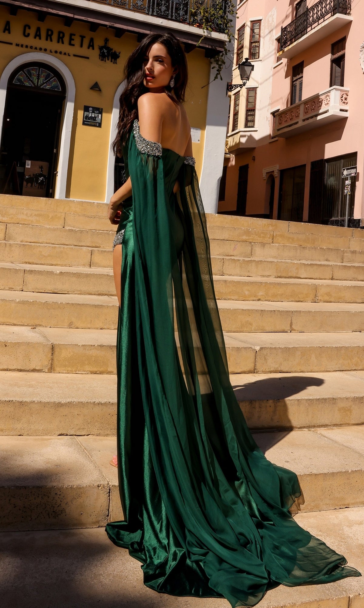 Wearing the Off-the-Shoulder Drapes Long Formal Dress R1312, a woman stands on outdoor steps and looks over her shoulder, with buildings and a restaurant entrance visible behind her.