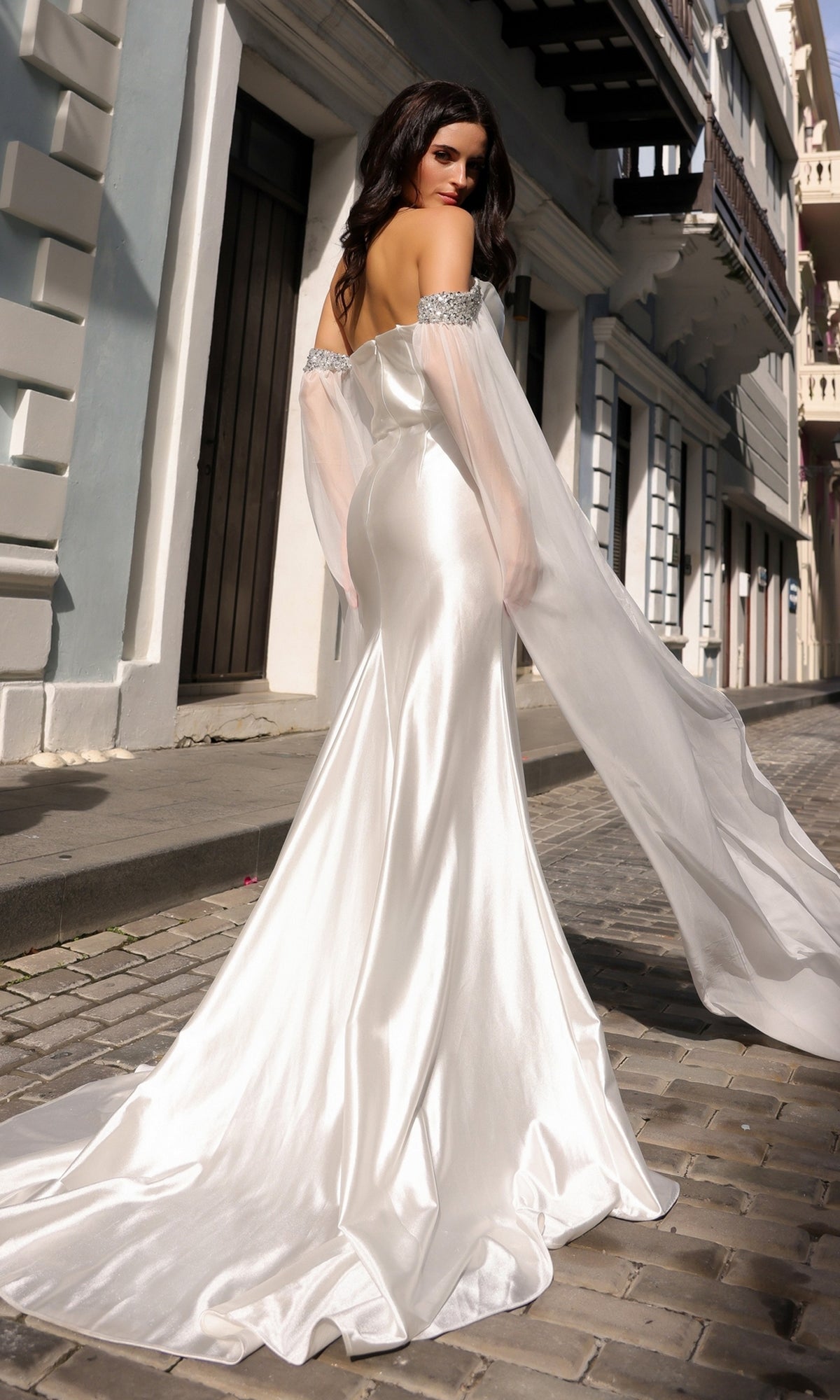 A woman wearing the Off-the-Shoulder Drapes Long Formal Dress R1312 with fitted style and draped sleeves stands on a cobblestone street, facing away from the camera.