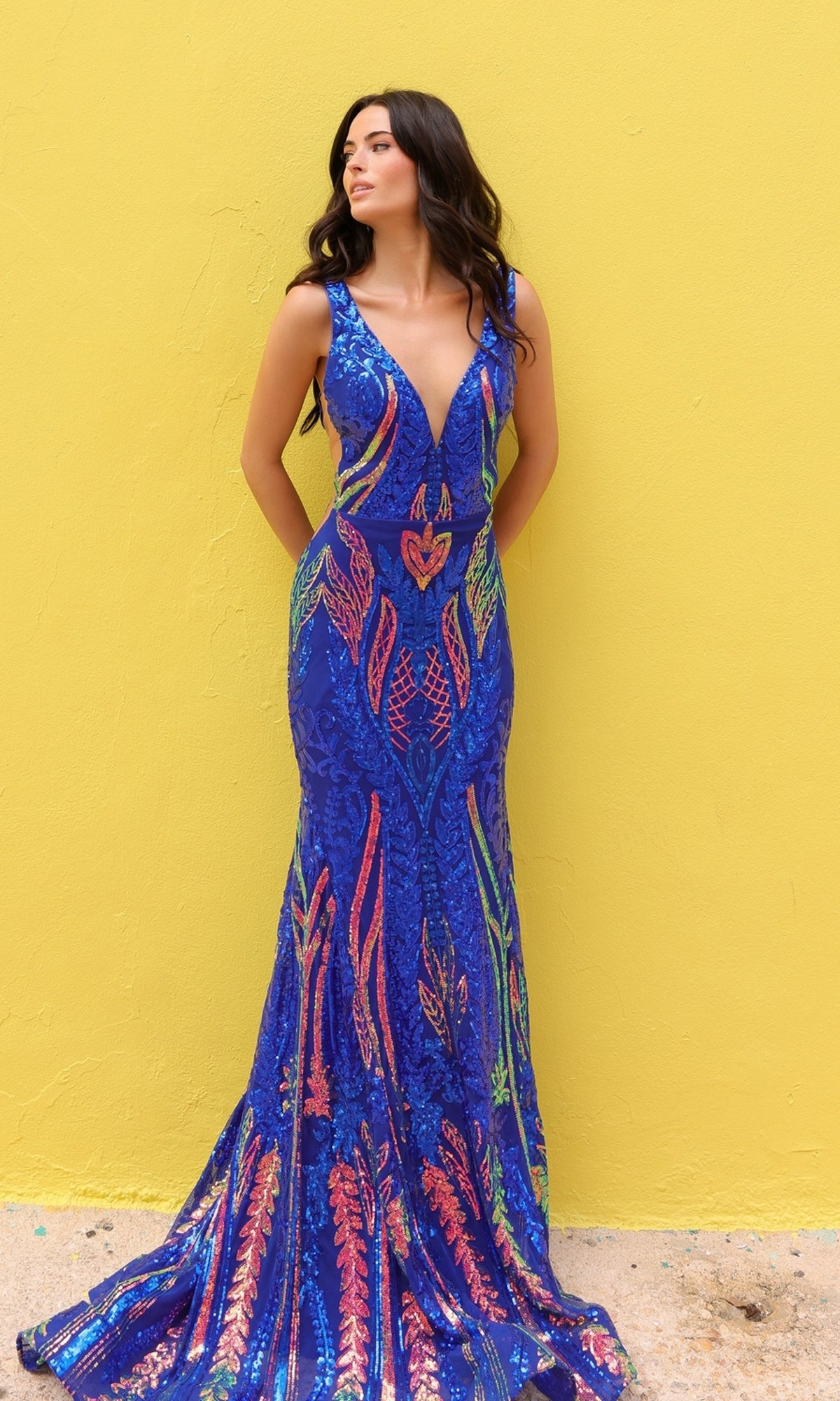 A woman wearing the Sheer-Sides Long Blue Sequin Prom Dress (R1402), featuring a sleeveless deep V-neck and multi-colored sequins, stands against a yellow wall, gazing to the side.
