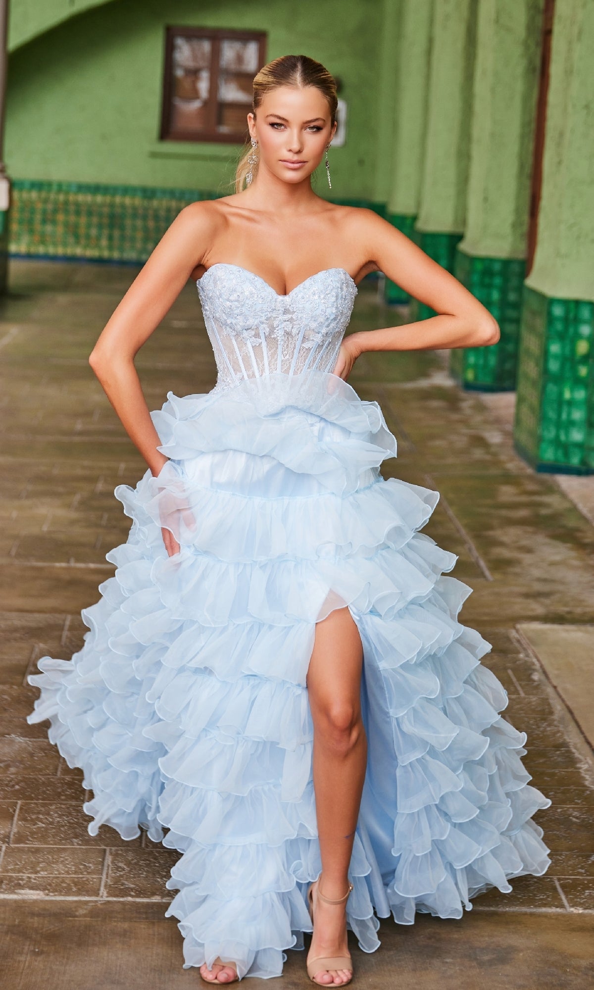 A woman wears the Nox Anabel R1688 strapless ruffled prom ball gown in light blue with a high slit, standing indoors on tiled floors with green walls and windows in the background.
