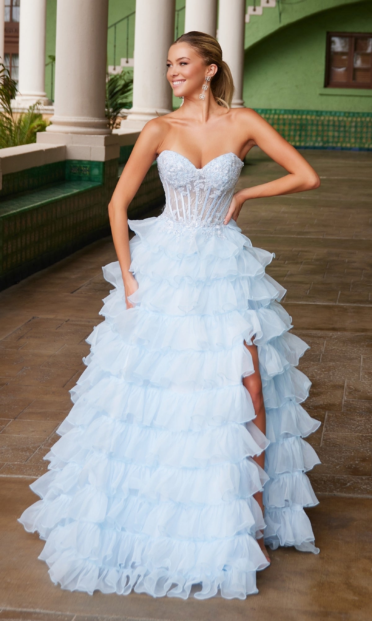 A woman stands outdoors, smiling with one hand on her hip, wearing the Strapless Ruffled Prom Ball Gown: Nox Anabel R1688, featuring a sheer corset bodice and thigh-high slit.