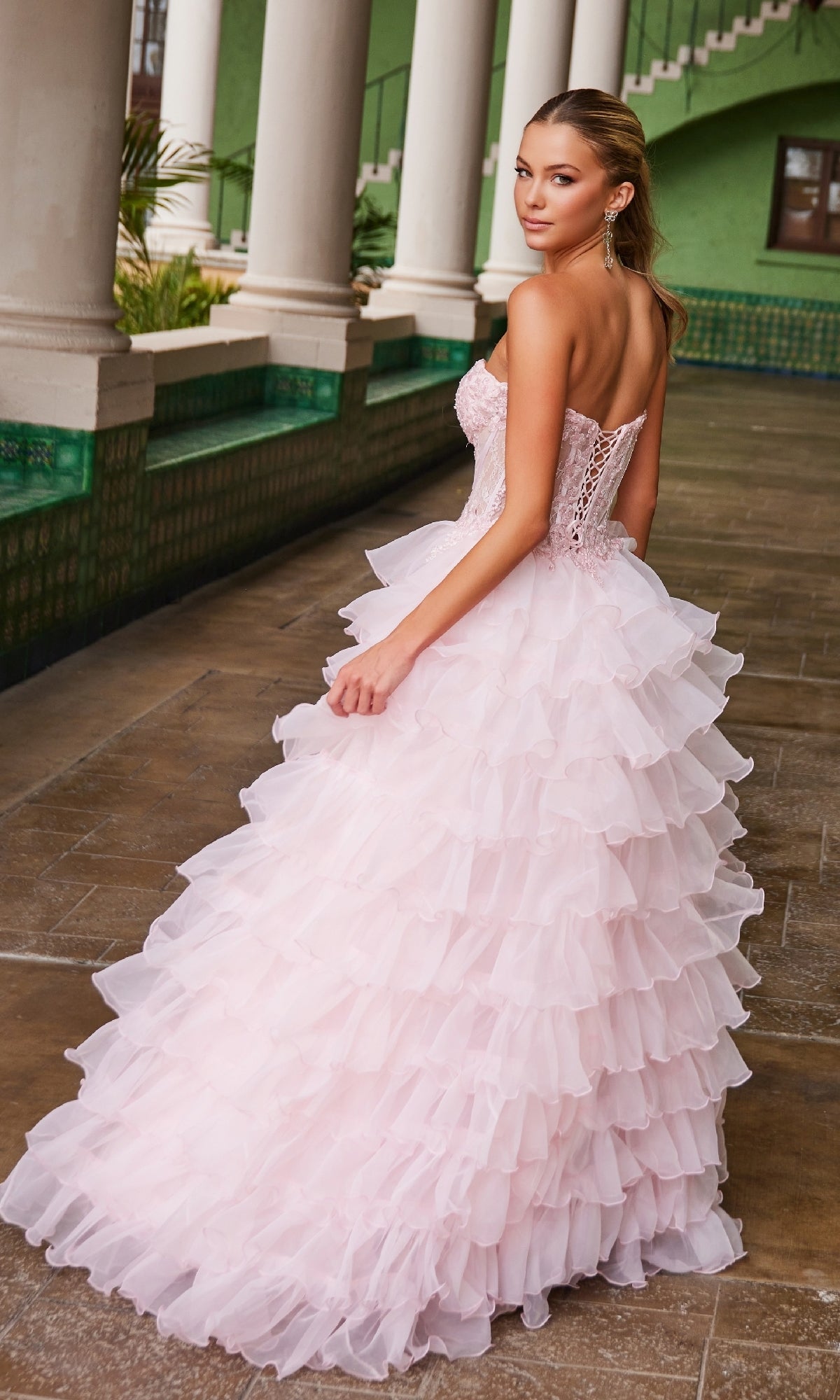 A woman wearing the Nox Anabel R1688 Strapless Ruffled Prom Ball Gown stands on a tiled walkway beside columns and a green building, looking over her shoulder.