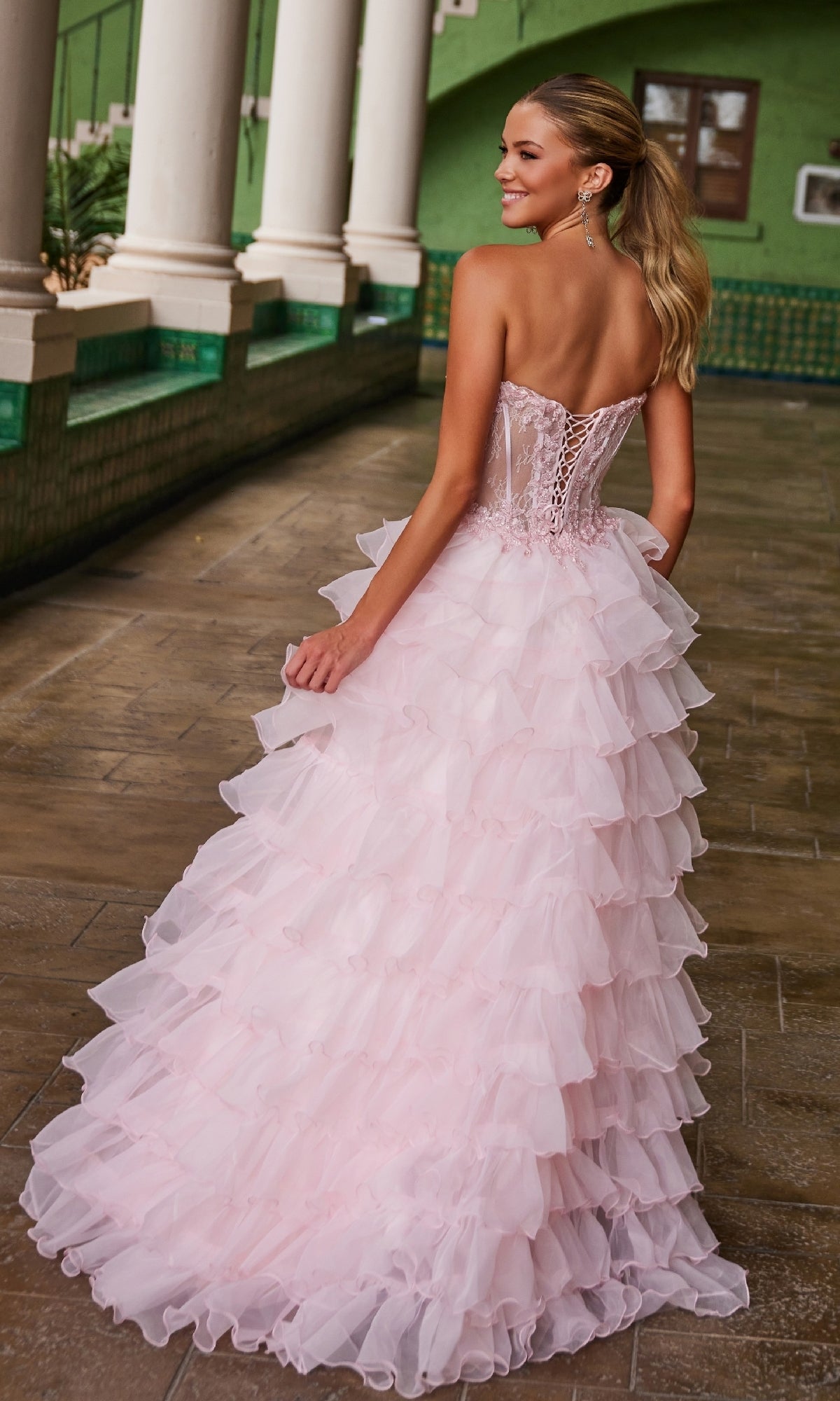 A woman in the Nox Anabel R1688 Strapless Ruffled Prom Ball Gown with a lace-up back stands on a tiled walkway near green walls and white columns, facing away from the camera.