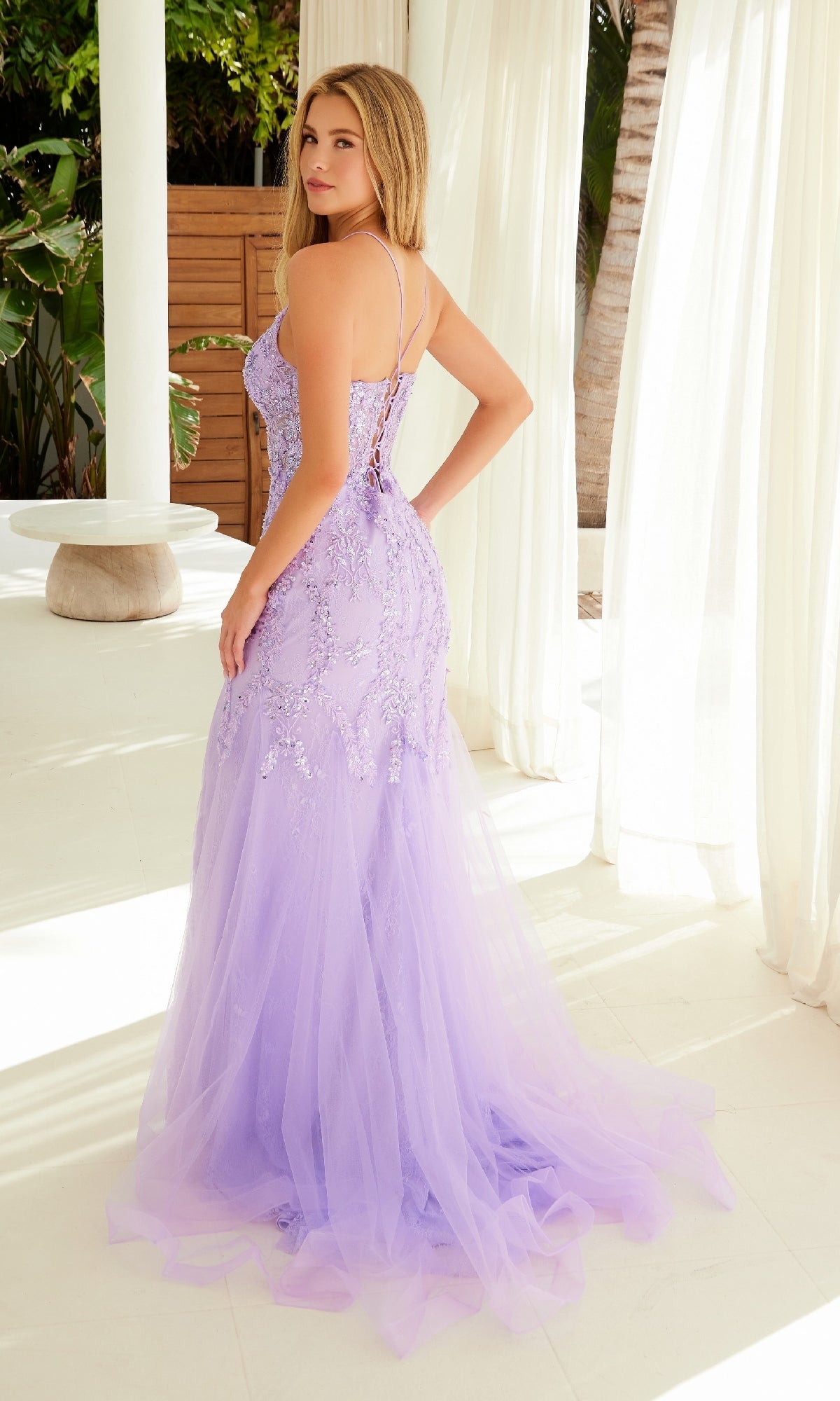 A woman stands indoors wearing the Nox Anabel R1717 Sequined Long Mermaid Prom Dress, featuring thin straps and a lace-up open back, as she looks over her shoulder at the camera.