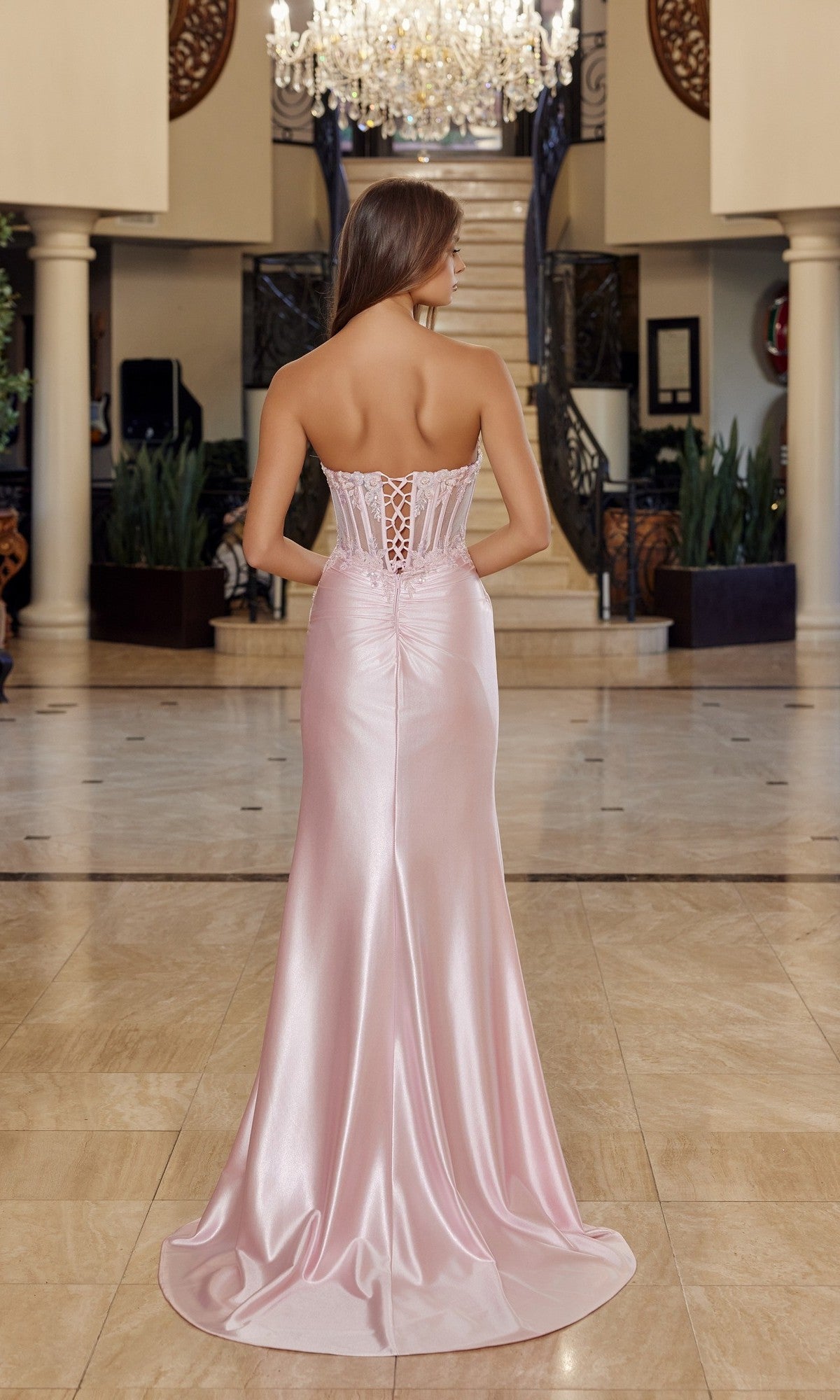 A woman wearing the Nox Anabel R1816 long prom dress, featuring a strapless satin design with a lace-up corset bodice, stands in an elegant marble-floored foyer, facing away from the camera.