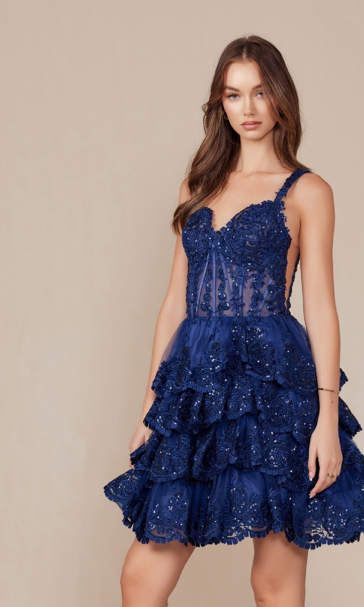 A woman showcases the Nox Anabel R880, a sleeveless navy blue lace cocktail dress with a fitted bodice and tiered ruffle skirt, standing against a plain beige background—the perfect short ruffled homecoming dress.