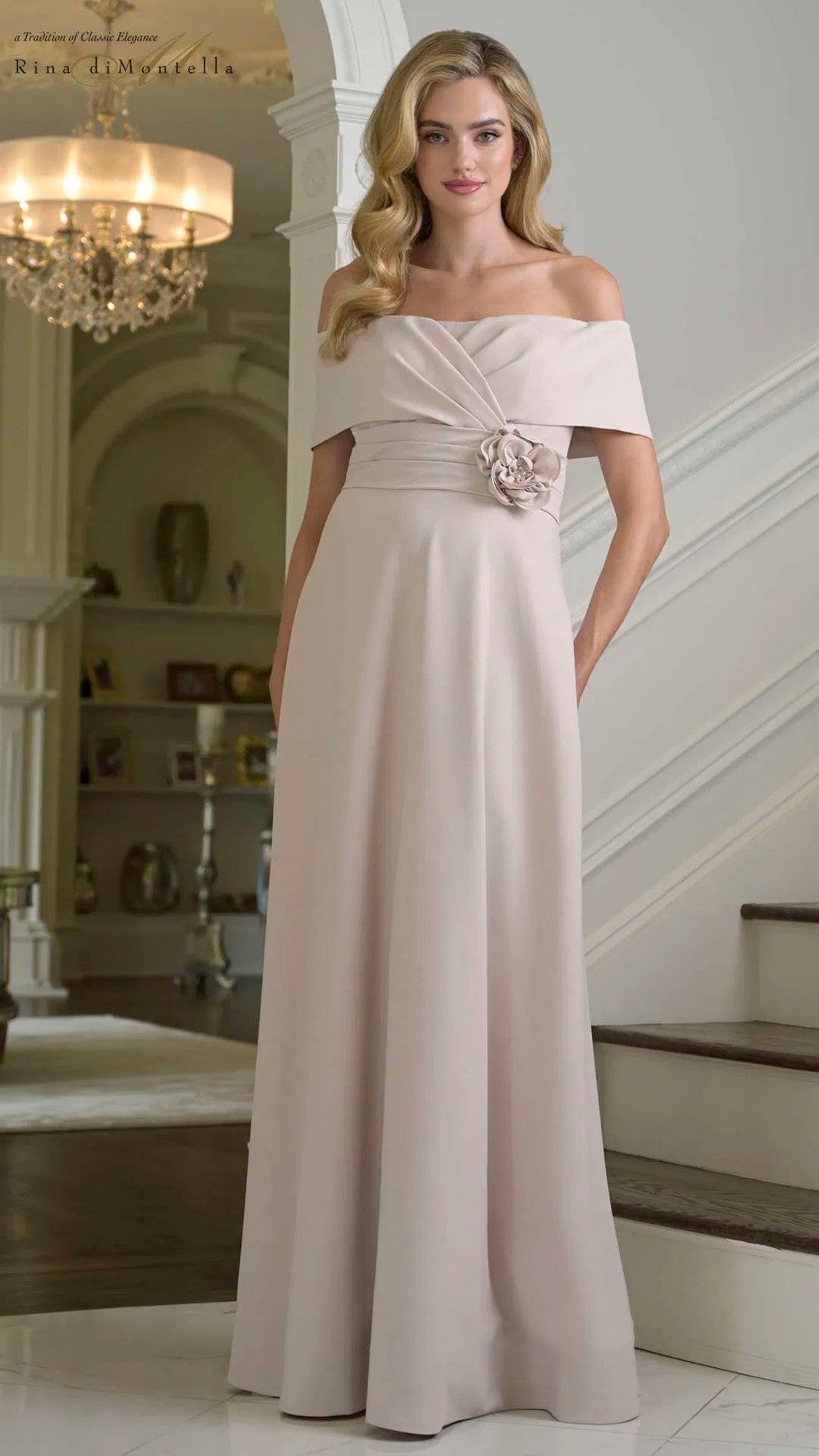 A woman with long, wavy hair poses near a staircase indoors wearing the Rina Di Montella RD3423, a champagne floor-length formal dress with an off-the-shoulder neckline, A-line skirt, and floral detail at the waist.