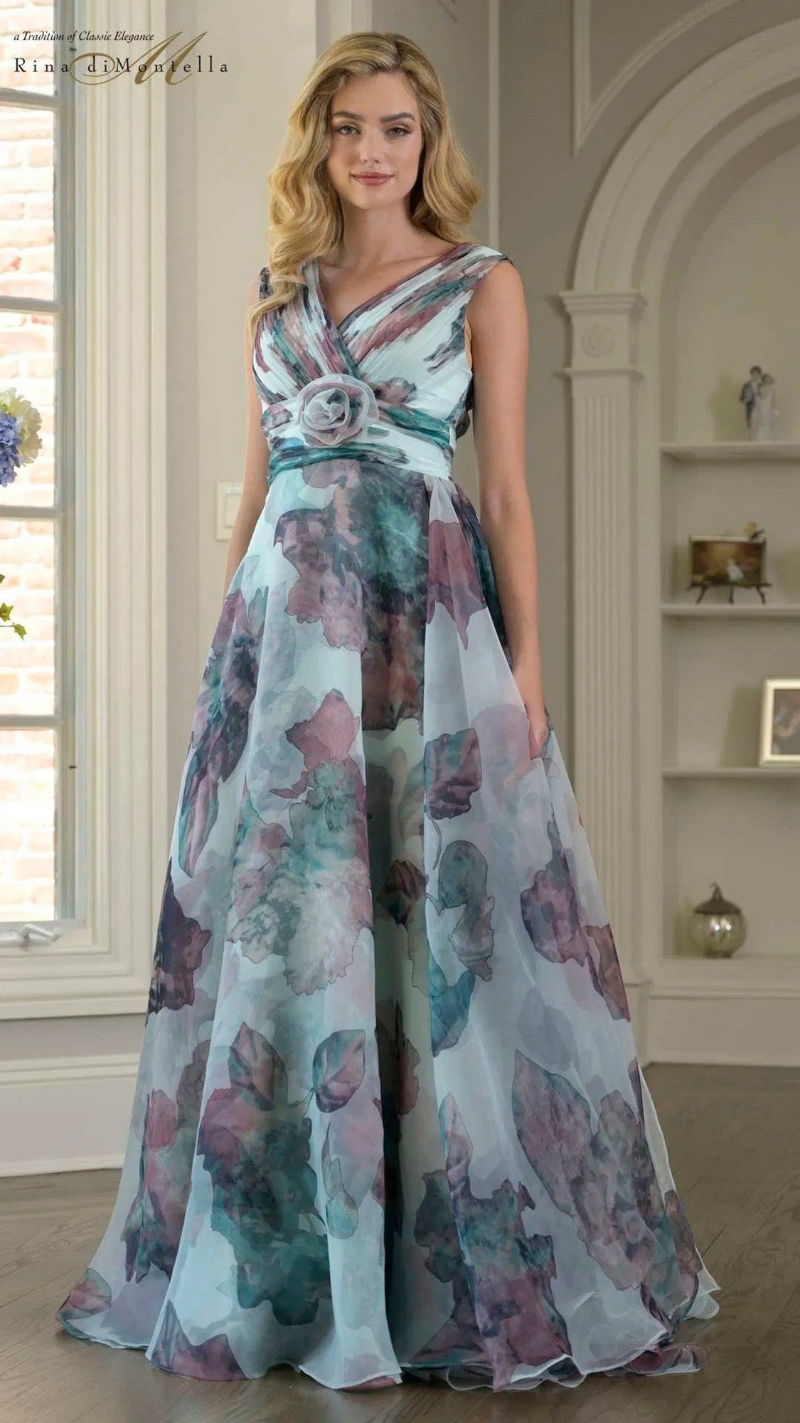A woman stands indoors wearing the Rina Di Montella RD3426 long formal dress in Tiffany Multi, featuring a floor-length organza design with a sleeveless cut, floral pattern, and rosette at the waist.