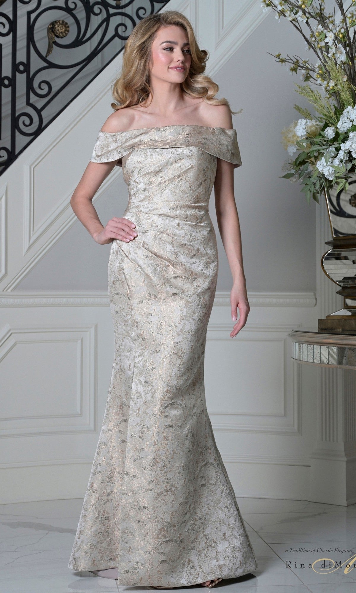 A woman in a champagne Long Formal Dress: Rina Di Montella RD3511 with an off-the-shoulder neckline and floor-length metallic brocade stands indoors beside a staircase and floral arrangement.