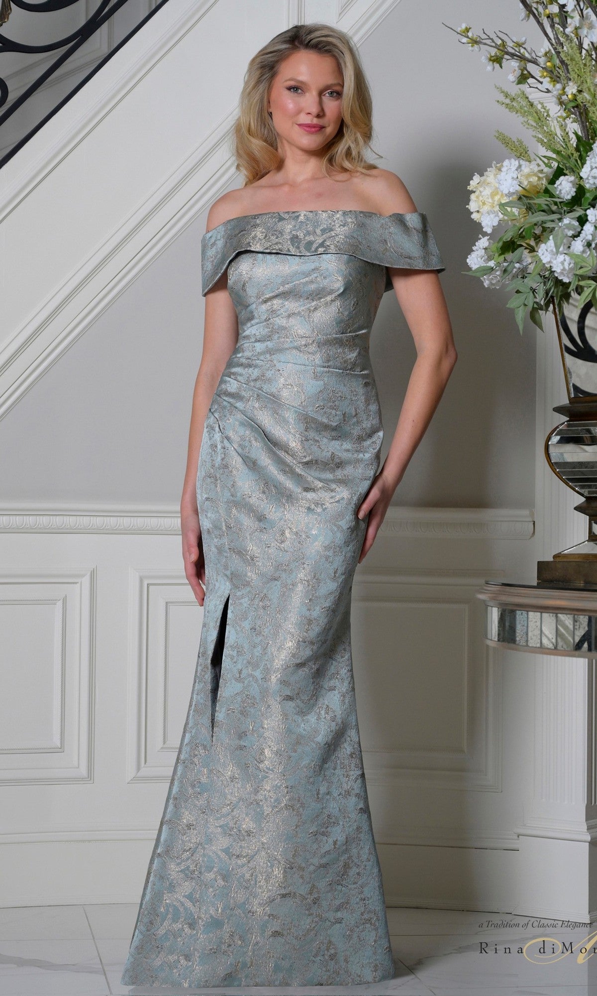 A woman in a deep sage Rina Di Montella RD3511 long formal dress with an off-the-shoulder neckline and front slit stands indoors near a staircase and floral arrangement.