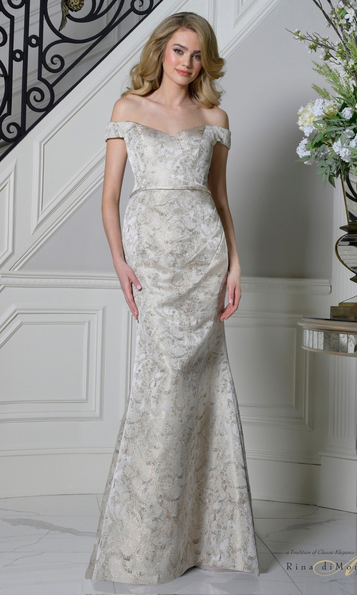A woman stands indoors in the Rina Di Montella RD3513, a champagne off-the-shoulder, floor-length formal dress with a fitted bodice and shimmering metallic jacquard—an elegant choice for the sophisticated mother of the bride.