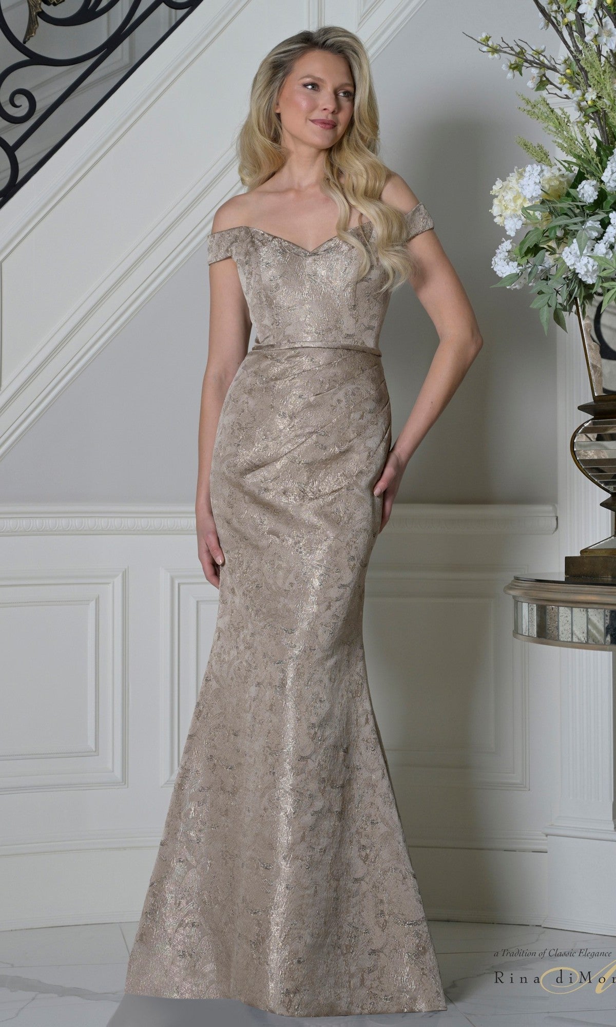 A woman in the Rina Di Montella RD3513 long formal dress in dark taupe, featuring a fitted bodice, jacquard fabric with subtle metallic pattern, off-the-shoulder neckline, and floor-length silhouette, stands by a staircase and elegant floral display—ideal for mother-of-the-bride.