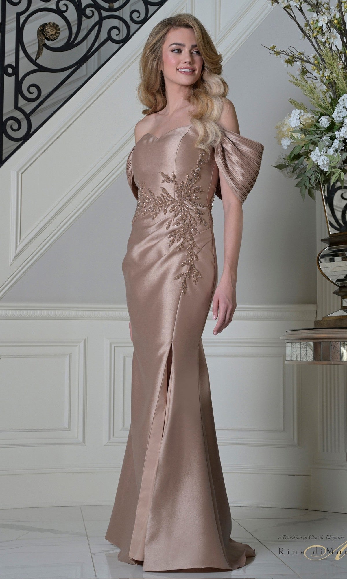 A woman stands indoors by a staircase, wearing the Rina Di Montella RD3527 long formal dress in rose gold, featuring off-the-shoulder pleated sleeves and elegant embroidery.