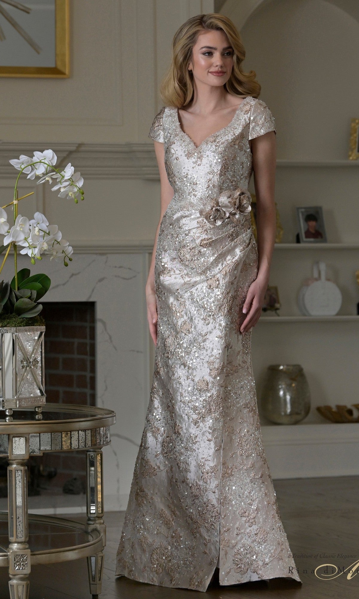 A woman in a floor-length, short-sleeve Rina Di Montella RD3529 gown in Gold Multi with metallic lace and floral details stands in a decorated living room near a table with a white orchid.