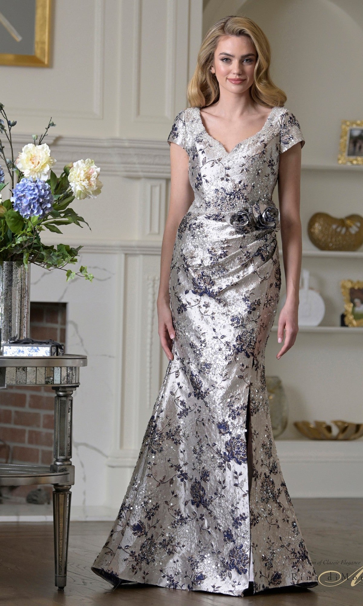 A woman stands indoors wearing the Long Formal Dress: Rina Di Montella RD3529 in Navy Multi, a floor-length mermaid gown with short sleeves and floral embellishments—perfect as a stunning mother-of-the-bride formal dress.