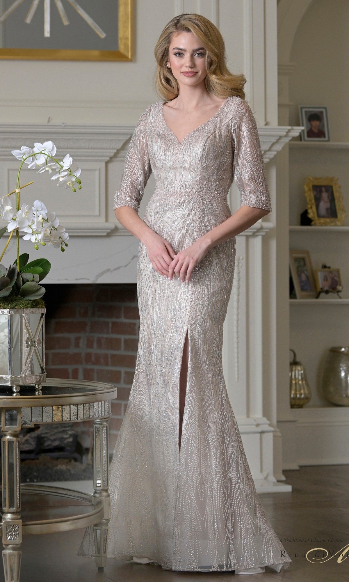 A woman poses indoors in front of a fireplace, wearing the Rina Di Montella RD3530 Long Formal Dress in light taupe—a floor-length, embroidered mesh mother-of-the-bride gown with three-quarter sleeves, v-neckline, and front slit.