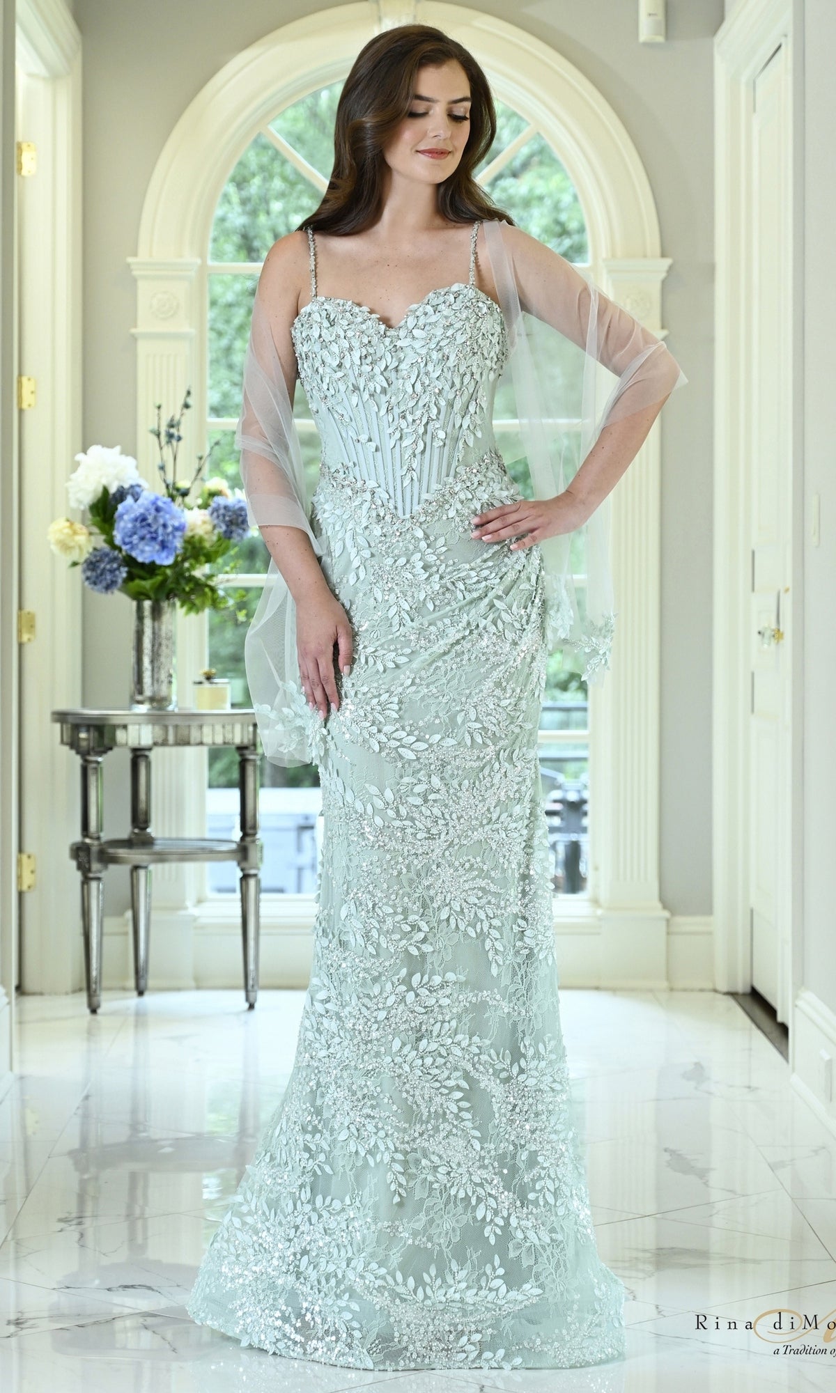 A woman wears a seaglass-colored Rina Di Montella RD3605 Corset Sweetheart Applique Formal Dress featuring a floor-length silhouette, leaf appliques, spaghetti straps, and a matching sheer shawl.
