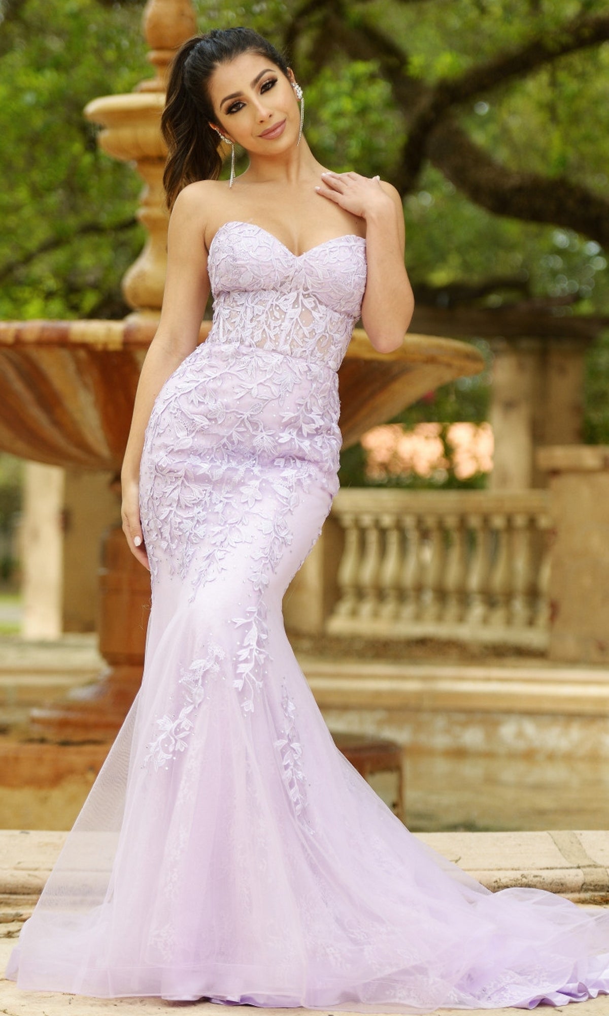 A woman poses outdoors in front of a fountain and trees wearing the Velvi Long Strapless Lace Mermaid Prom Dress Reva, a fitted lavender gown.