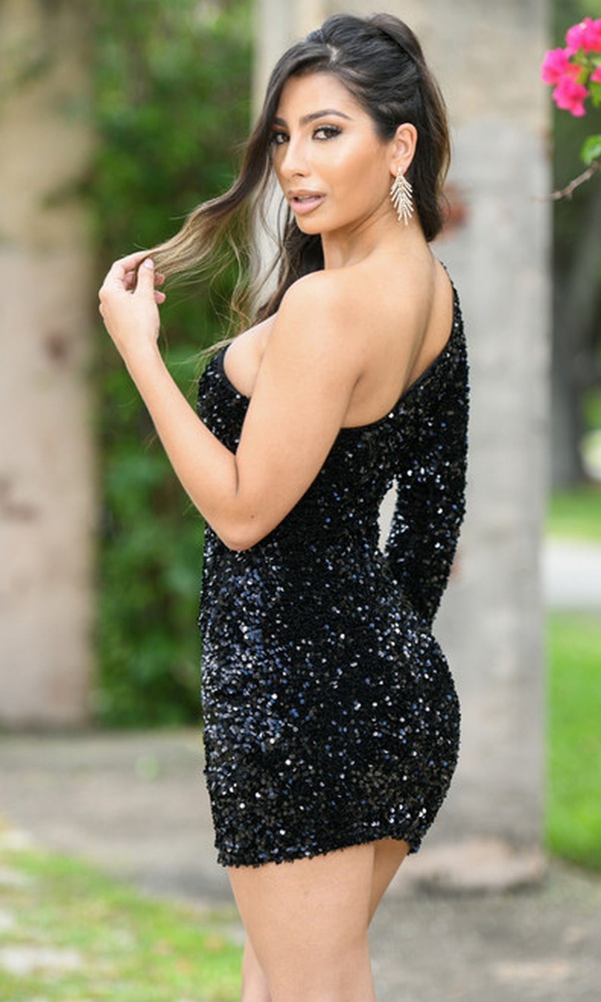 A woman stands outdoors in the Rita One-Sleeve Short Sequin Homecoming Dress with statement earrings, looking over her shoulder at the camera.