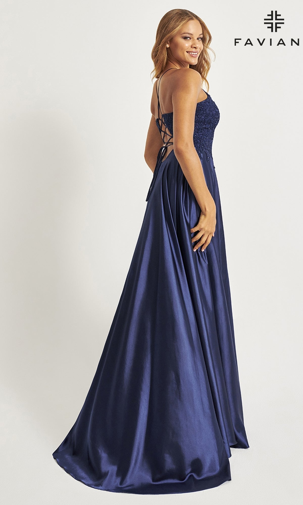 A woman stands and smiles against a plain light background, wearing the Faviana Lace-Bodice Long A-Line Prom Dress in navy blue satin with a fitted lace-up back and delicate embroidered lace details.