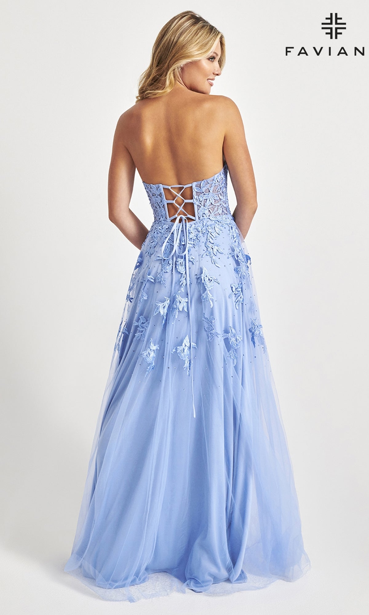 A woman faces away from the camera in the Faviana Strapless Sweetheart Long Lace Prom Dress, featuring a light blue hue, lace-up back, and floral embroidery, standing against a plain background.