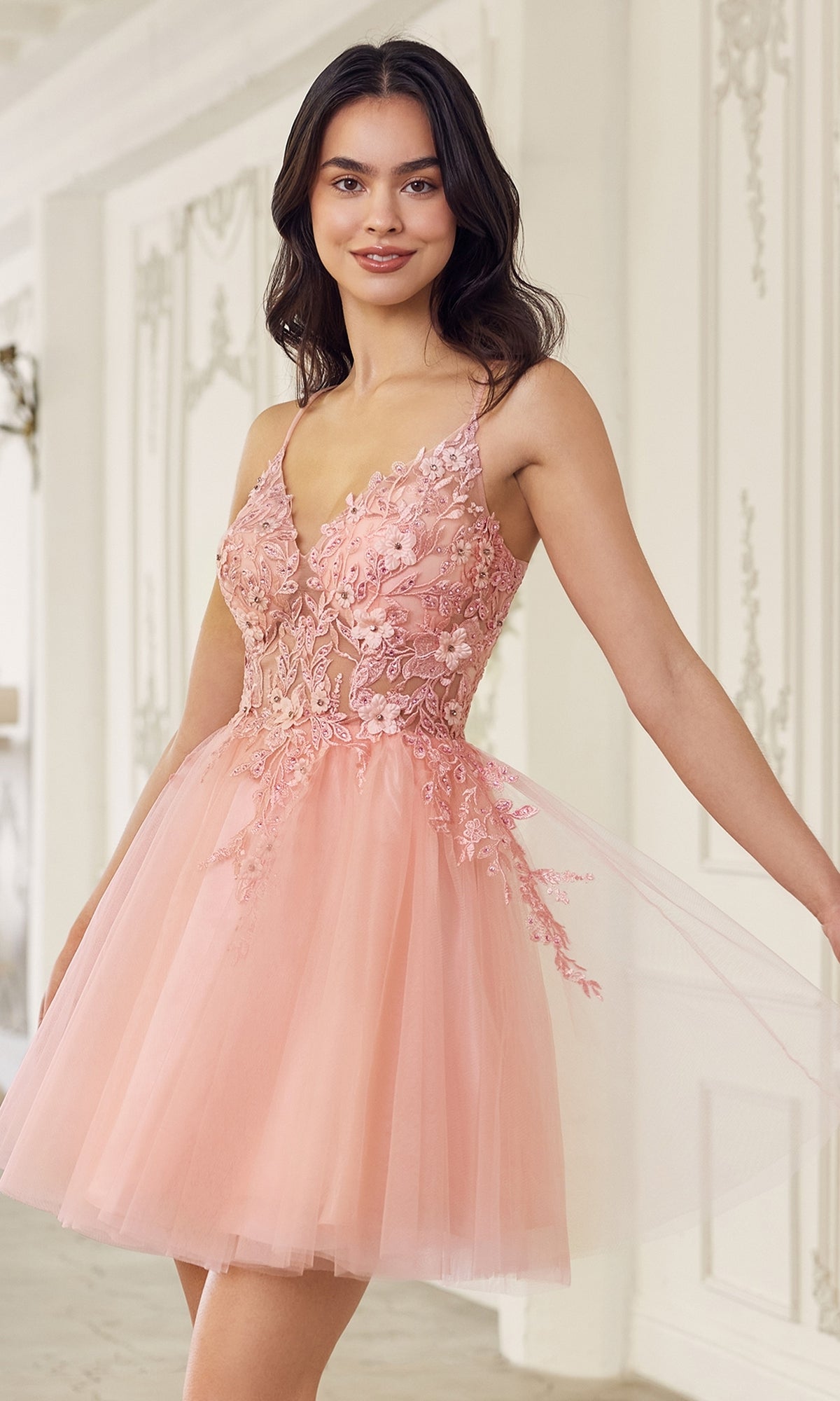 A woman stands indoors, smiling in the Short Babydoll Homecoming Dress: CLSF047—an A-line party dress with a sleeveless embroidered lace bodice and a knee-length pink tulle skirt.