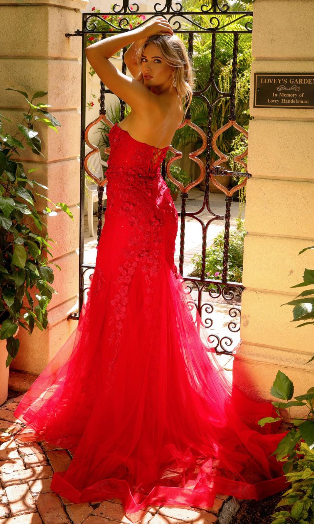 A woman in the Amelia Couture Corset Mermaid Prom Dress SU081 stands before a gated garden entrance, surrounded by lush greenery and sunlight.