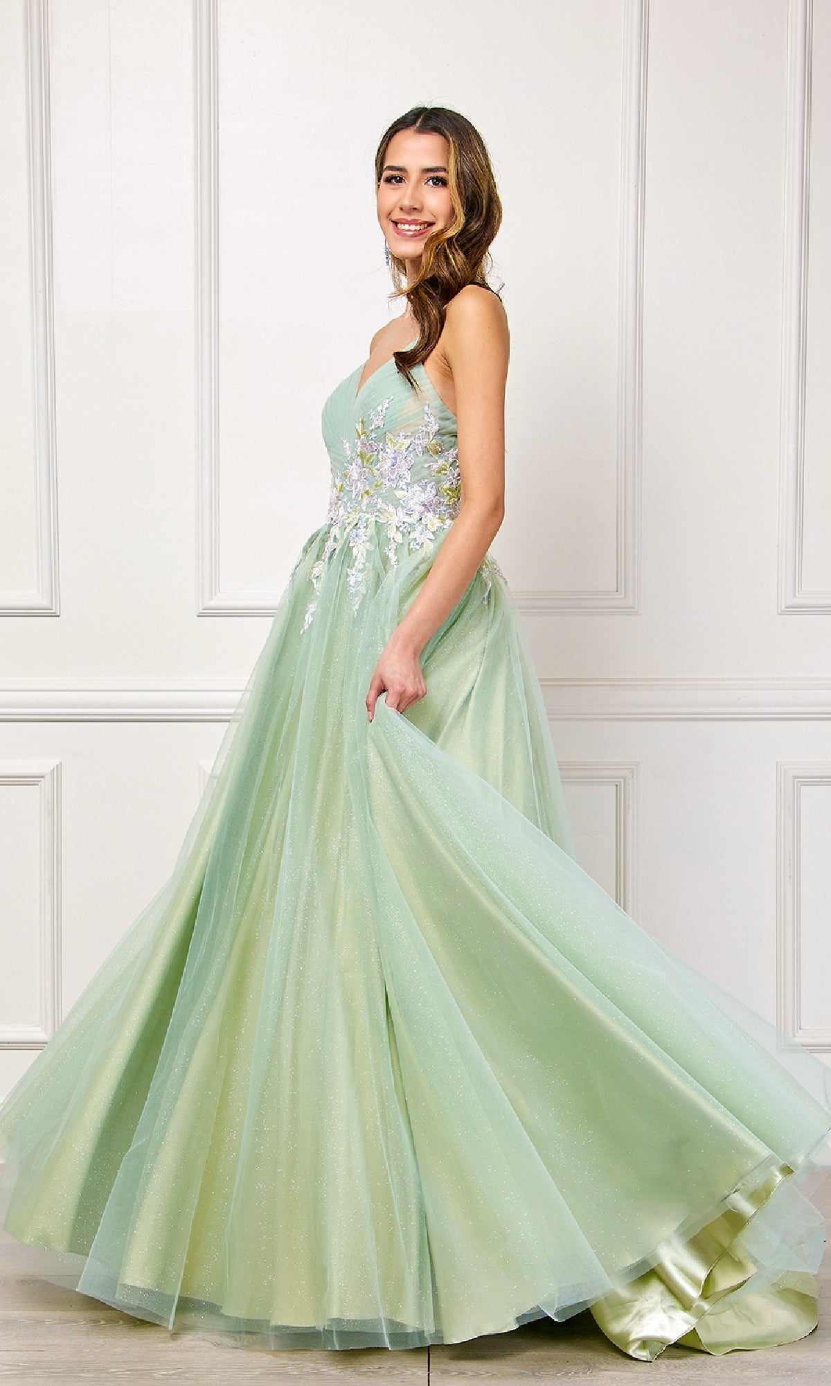 A woman wearing the Long Pastel Prom Ball Gown: Amelia Couture SU088, featuring floral embroidery on the bodice, stands before a white paneled wall and gracefully holds her gown.
