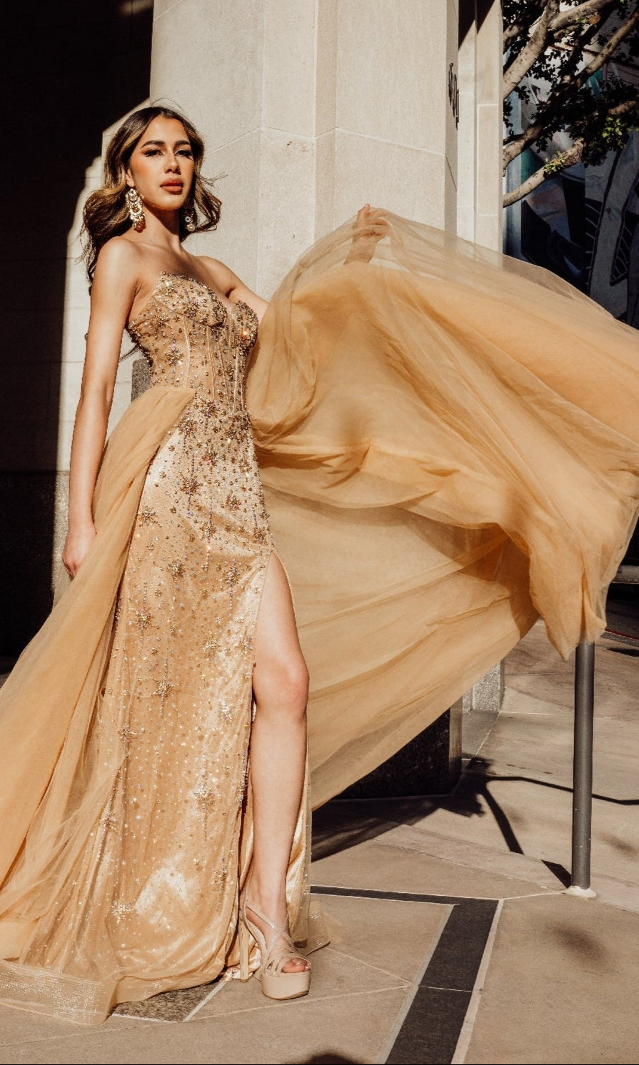 A woman models the Mac Duggal SU091, a strapless champagne formal dress, outdoors. She lifts the gown’s flowing fabric to highlight its elegant beaded details and a thigh-high slit.