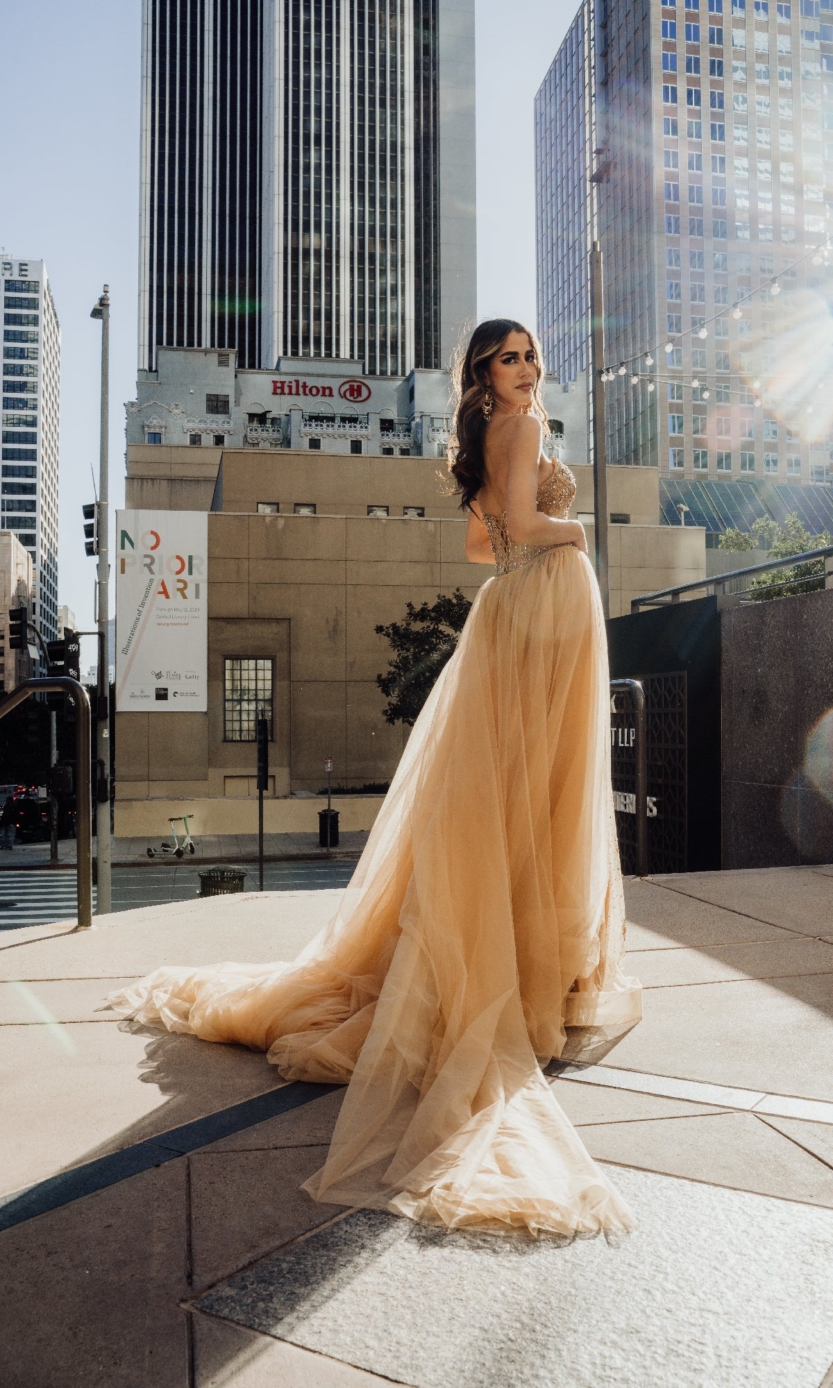 A woman in the Strapless Champagne Formal Dress: Mac Duggal SU091 stands outdoors on city steps, with tall buildings and sunlight in the background.