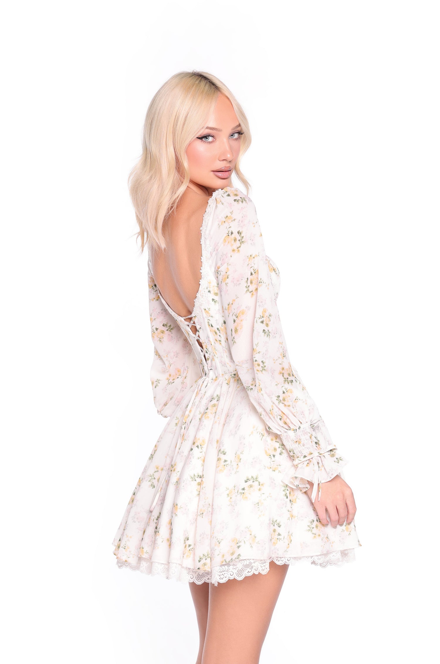A woman wears the Sadie Yellow Floral Short Casual Dress, featuring a buttercream hue and lace-up back, as she stands looking over her shoulder against a white background.