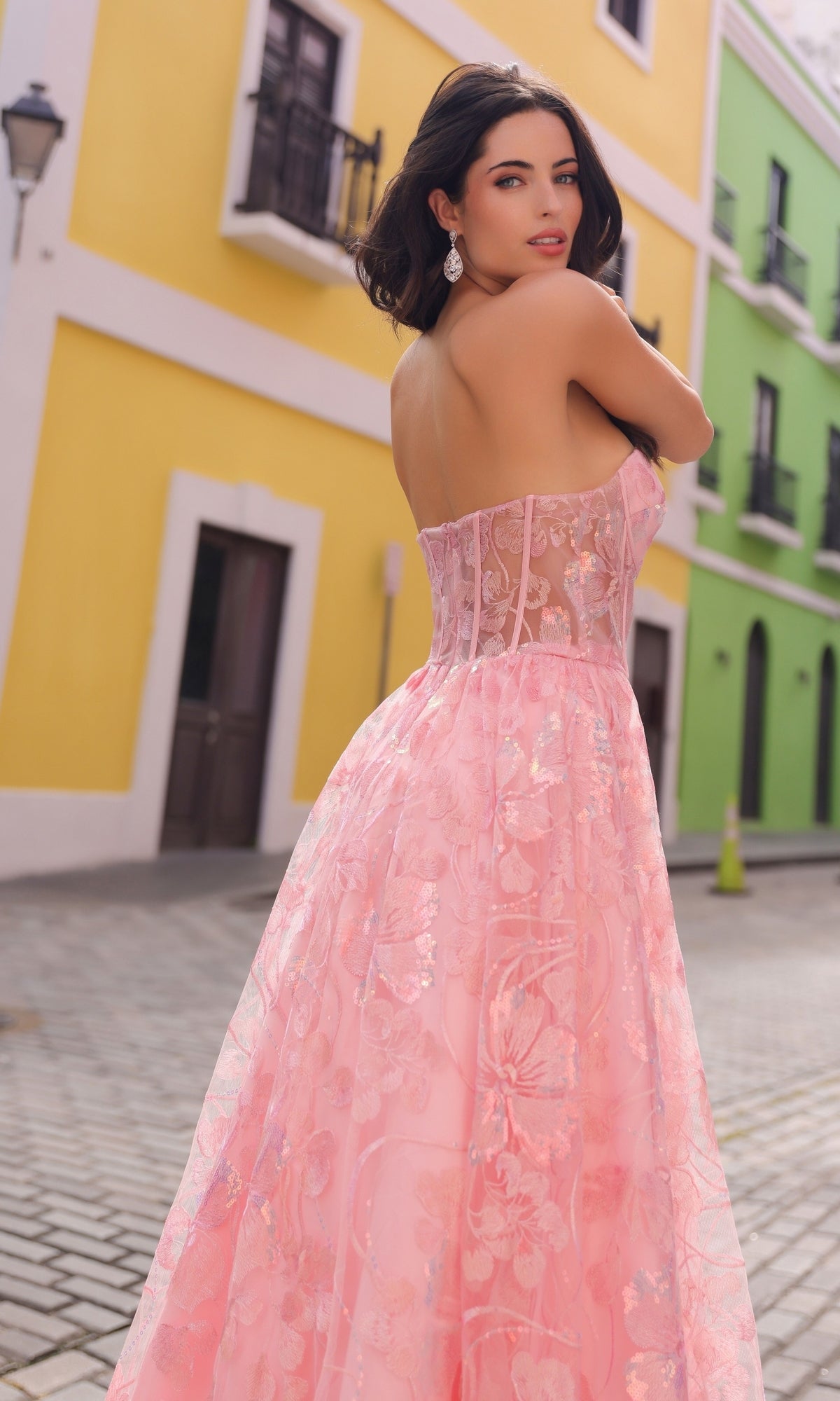 Wearing the Sequin-Floral Strapless Long Prom Ball Gown T1332, a woman stands on a cobblestone street with colorful buildings, glancing over her shoulder.