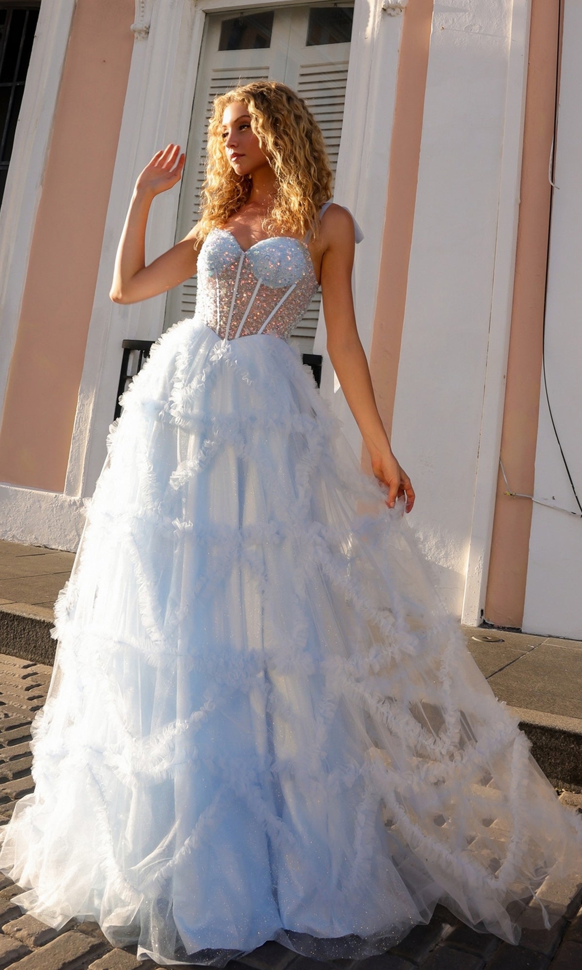A woman stands outside a pale pink and white building wearing the Shoulder-Bows Long Ruffled Prom Gown T1340—a sheer, light blue tulle dress with a beaded corset bodice and full skirt.