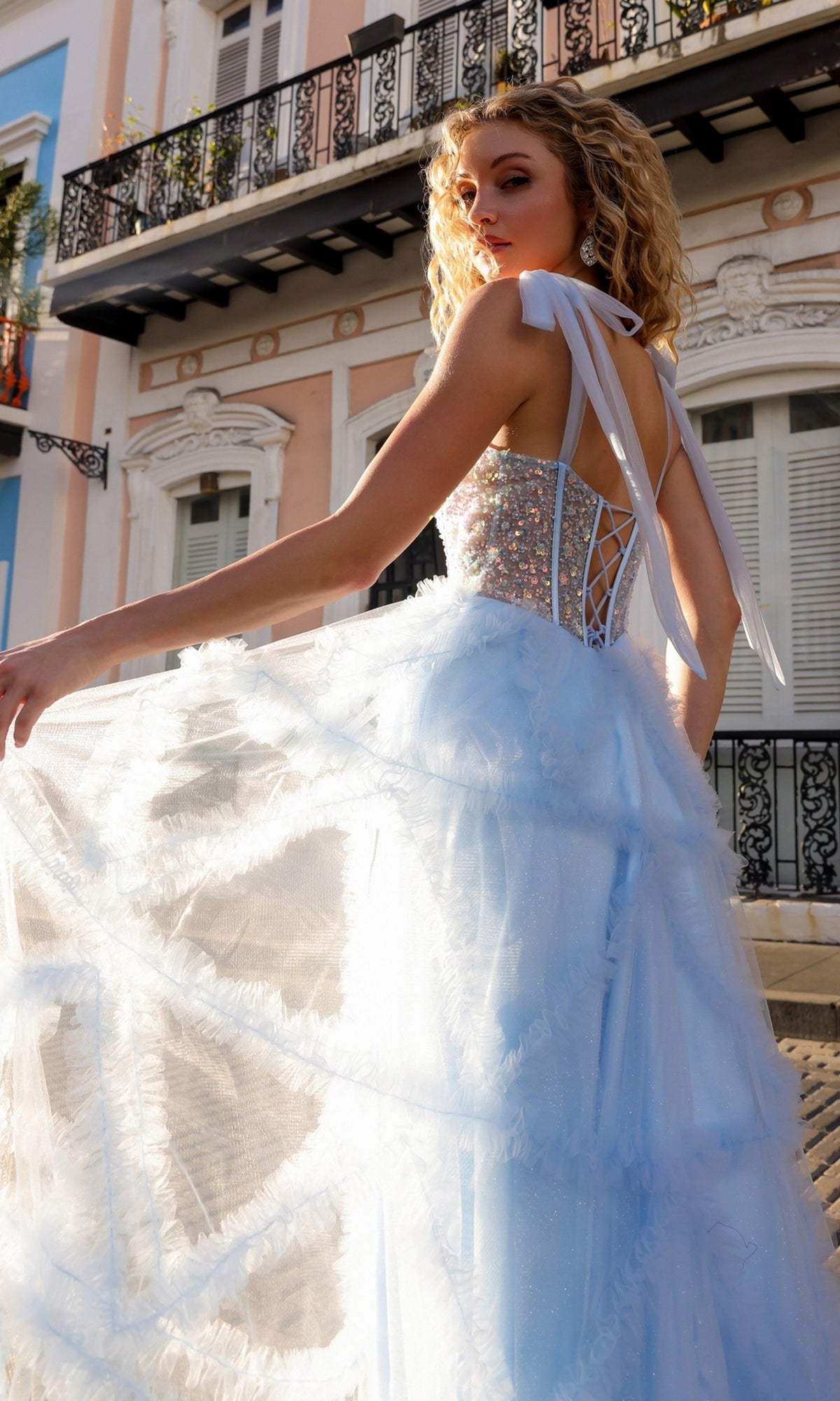 A woman stands outdoors before pastel buildings, gracefully holding her Shoulder-Bows Long Ruffled Prom Gown T1340, featuring a light blue tulle design and a beaded corset bodice.