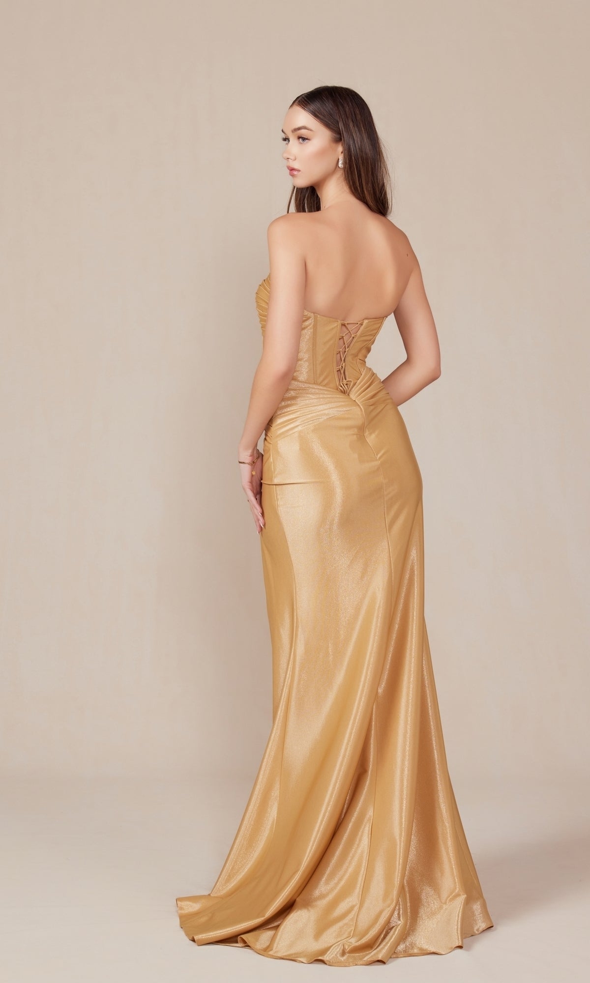 A woman faces away in a neutral studio, wearing the Nox Anabel T1489 Strapless Lace-Up Long Prom Gown with a metallic gold finish, fitted bodice, and lace-up back.