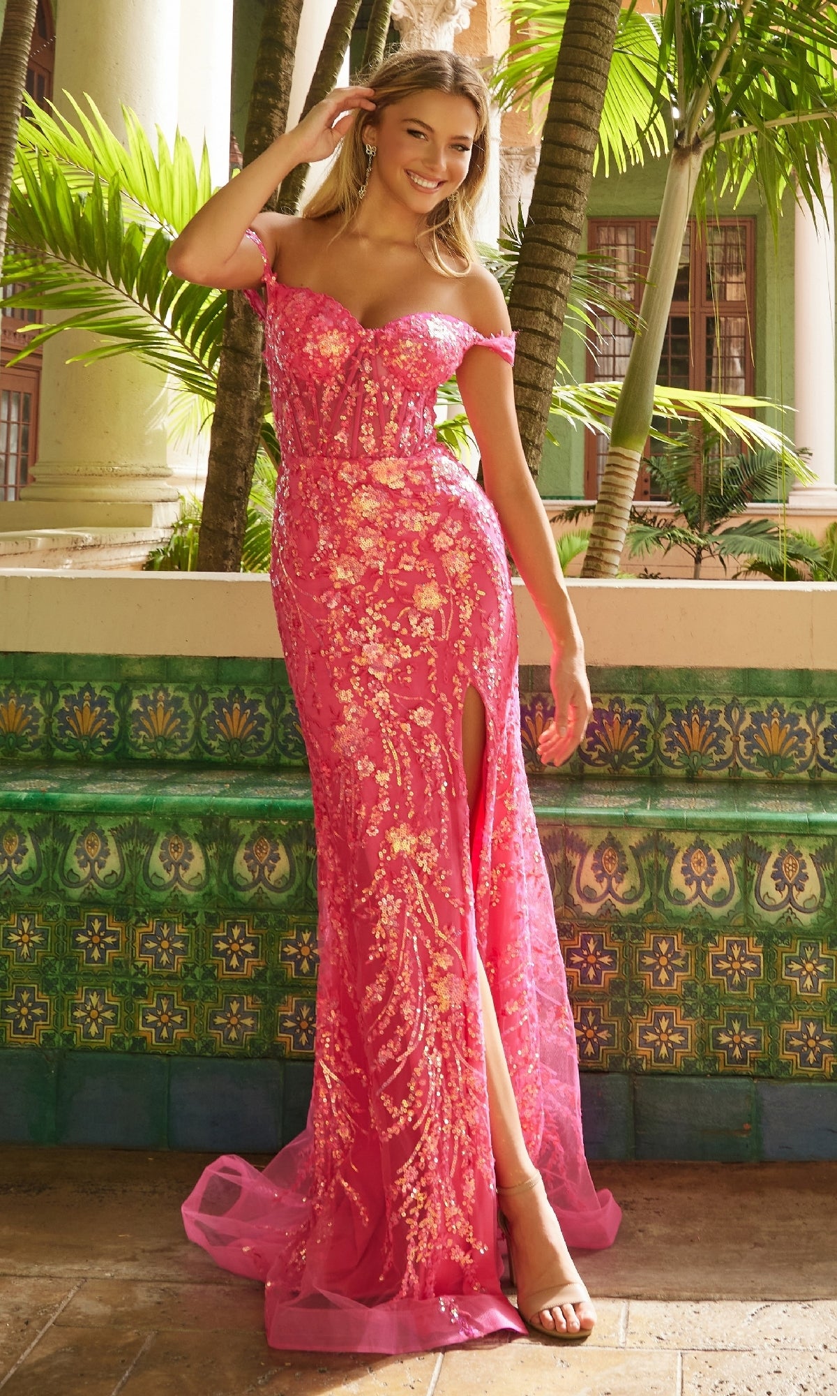 A woman stands outdoors in the Nox Anabel T1504 long sequin-print prom dress, featuring pink off-the-shoulder styling and a thigh-high slit. Green tiled walls and palm plants in the background enhance the elegant look.