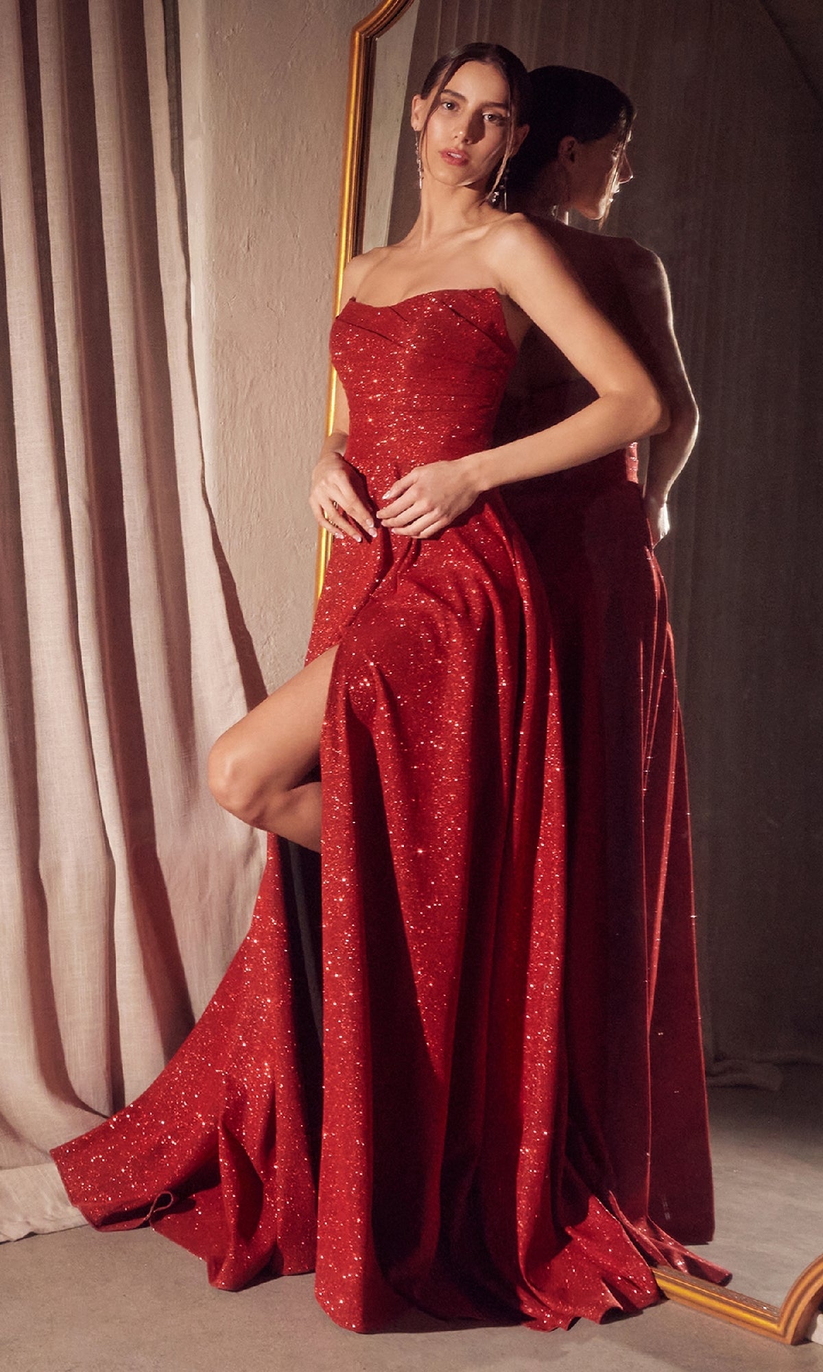 A woman in the Amoris T701 strapless long A-line glitter prom dress with a high slit stands by a mirror, her reflection shimmering in the sparkly red gown.
