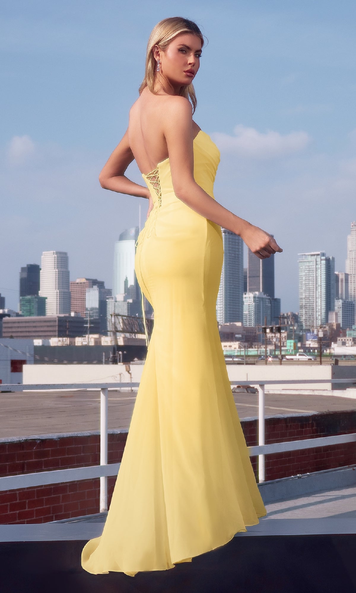 A woman in the Lace-Up Strapless Prom Gown: Amoris T702 stands on a rooftop, her fitted yellow dress glowing against the city skyline.