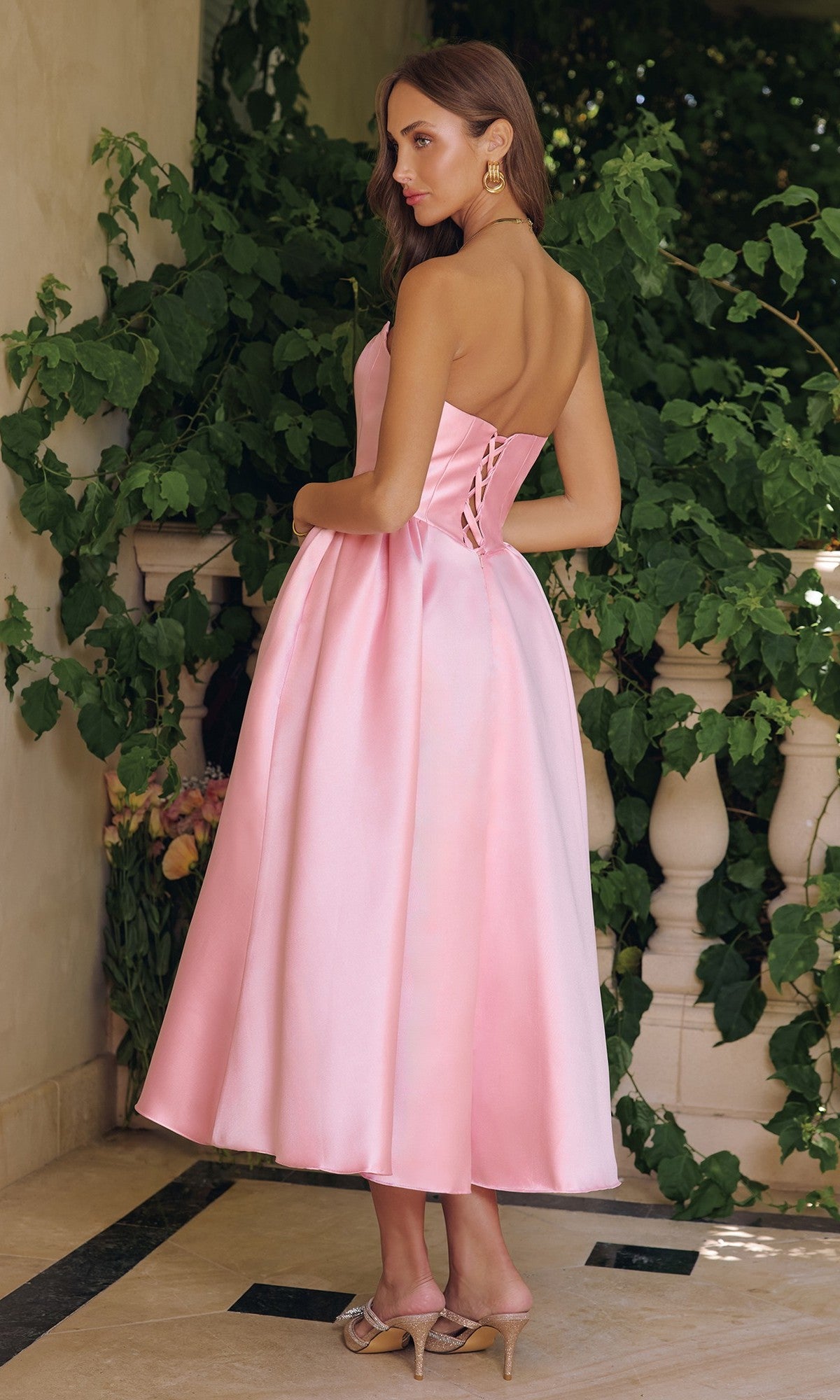 A woman stands on a patio wearing the Amoris T822 Midi Prom Dress with an A-line skirt and lace-up back, paired with open-toe heels. Green foliage and a white railing complete the backdrop.