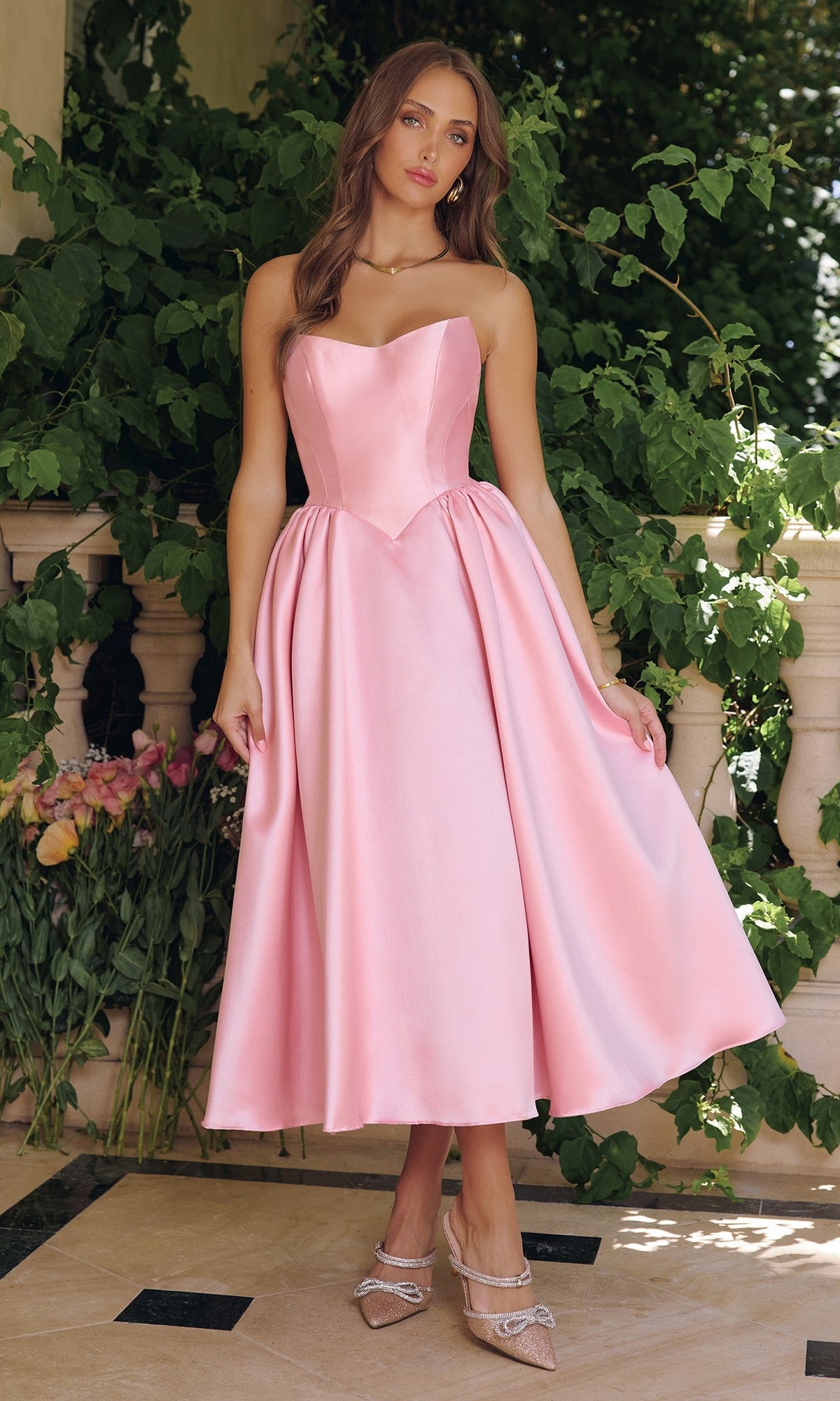 A woman stands outdoors in a strapless Midi Prom Dress: Amoris T822 in rose pink with an a-line, flared silhouette and light pink heels; green foliage and flowers are seen in the background.