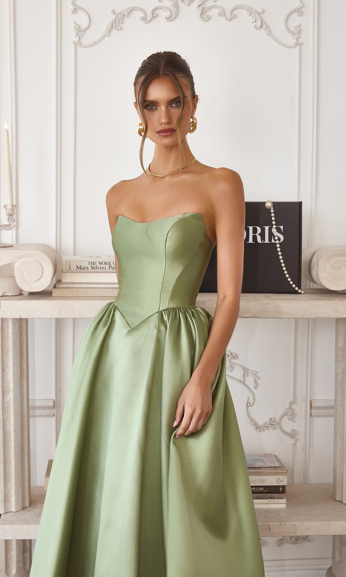 A woman wearing the Amoris T822 Midi Prom Dress with a strapless bodice and elegant A-line skirt stands indoors before shelves lined with books and decor.